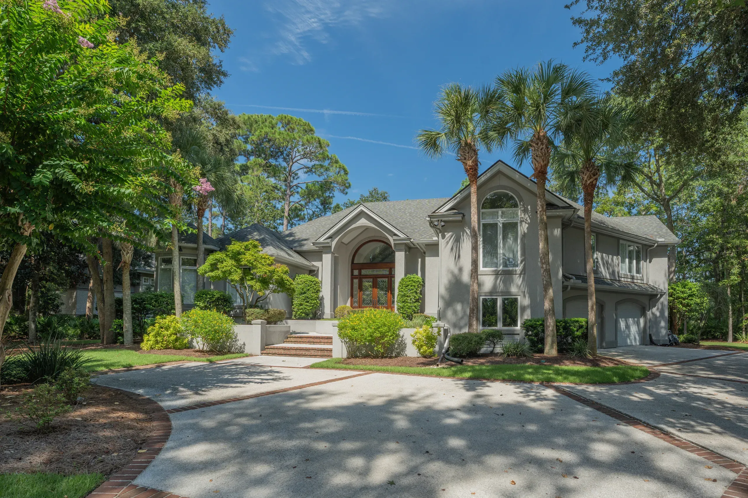 A two-story stucco home with a curved arched entry and tall windows sits behind a circular driveway shaded by mature palm and oak trees.