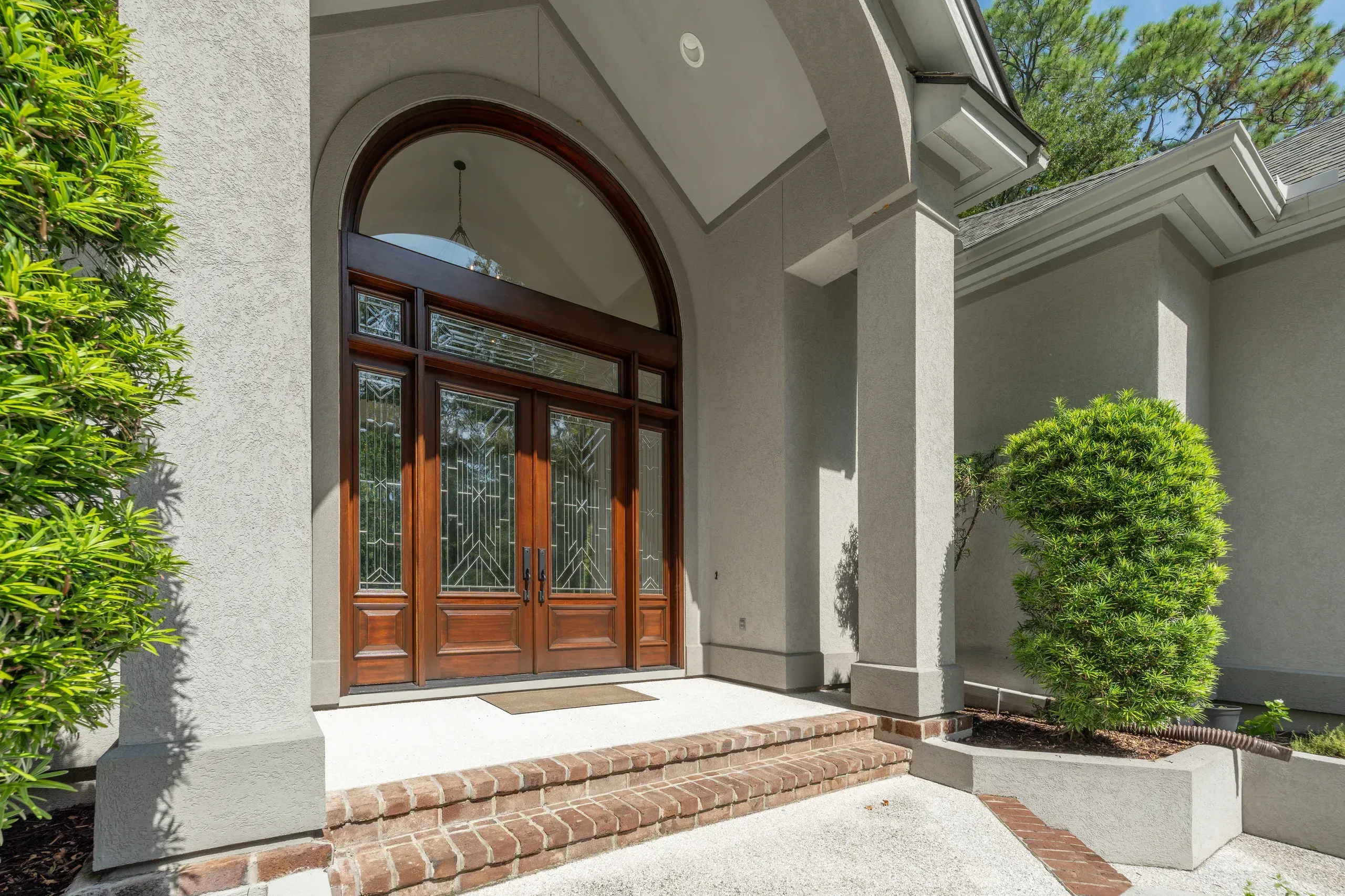 An arched stucco entryway shelters a stained wood double door with decorative glass panels, approached by brick steps and flanked by lush green shrubs.
