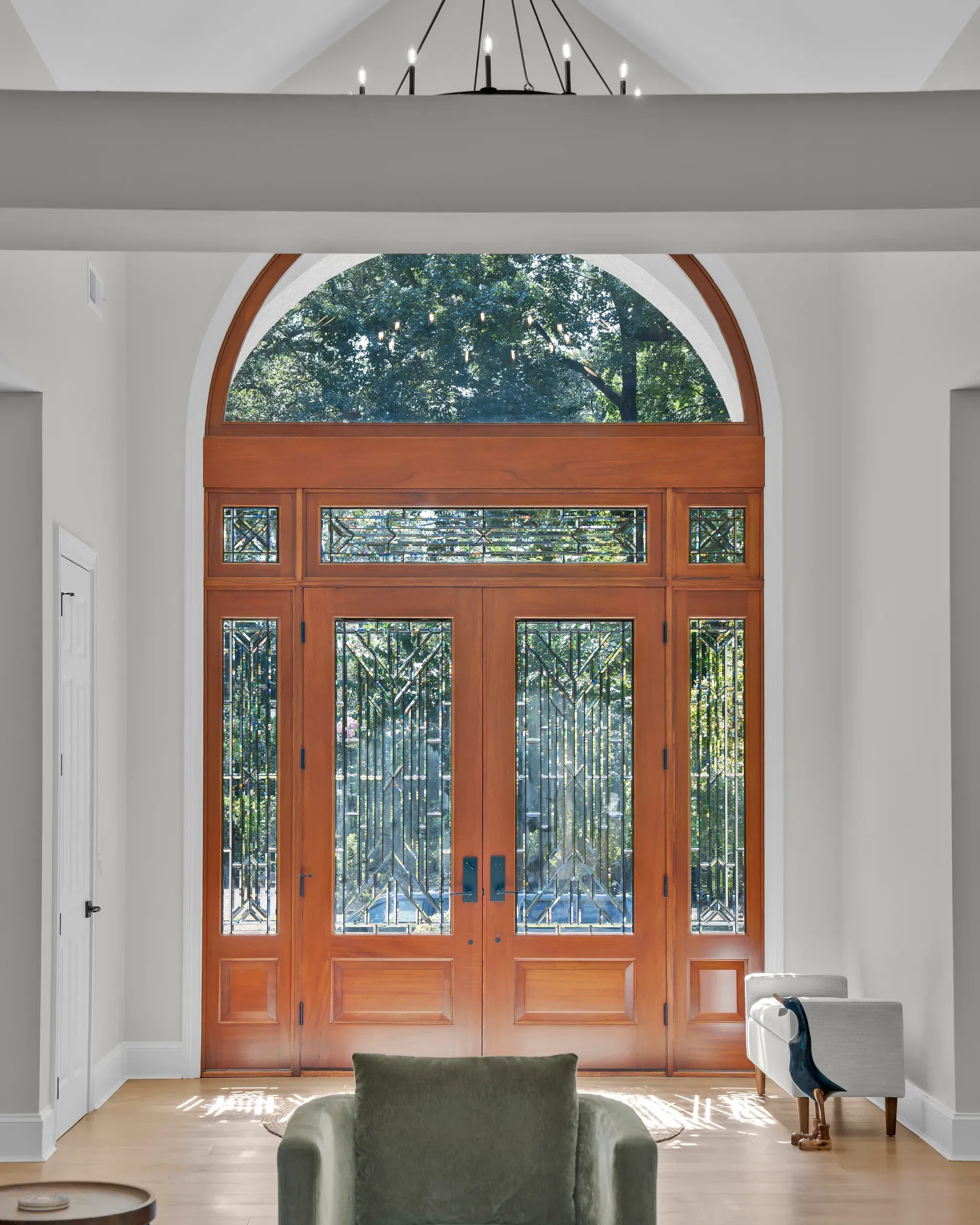 A grand wood front entry with double doors featuring geometric stained glass panels and an arched transom window overlooking a leafy tree canopy, framed by a cathedral ceiling and modern chandelier.