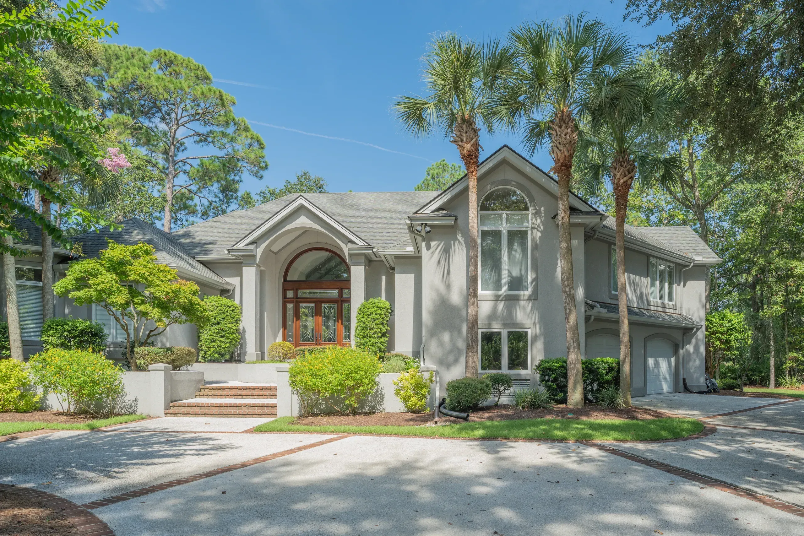A two-story stucco home with an arched wood-and-glass entryway is framed by tall palm trees and mature pines along a curved brick-edged driveway.