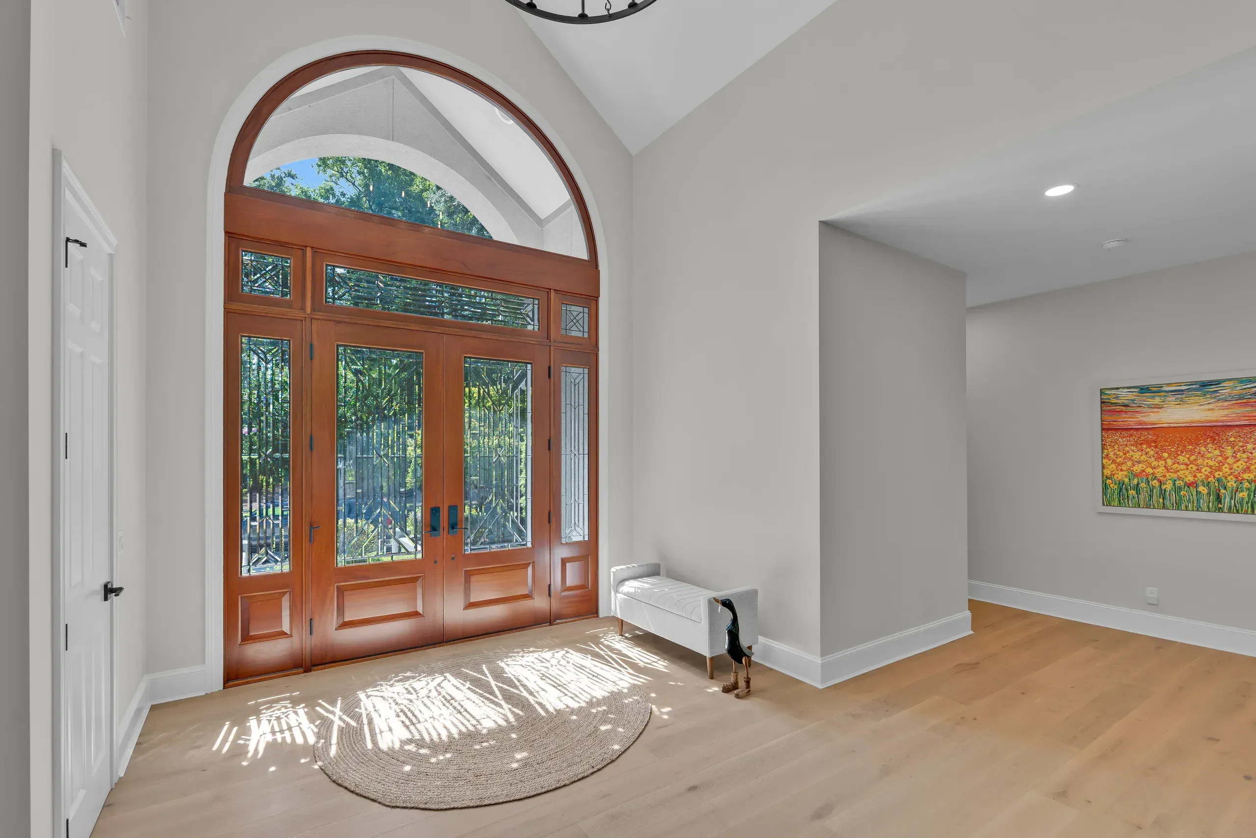 A double-height entryway features a stained wood double front door with decorative glass panels and an arched transom window, opening onto a light oak floor with a round jute rug and an upholstered bench.