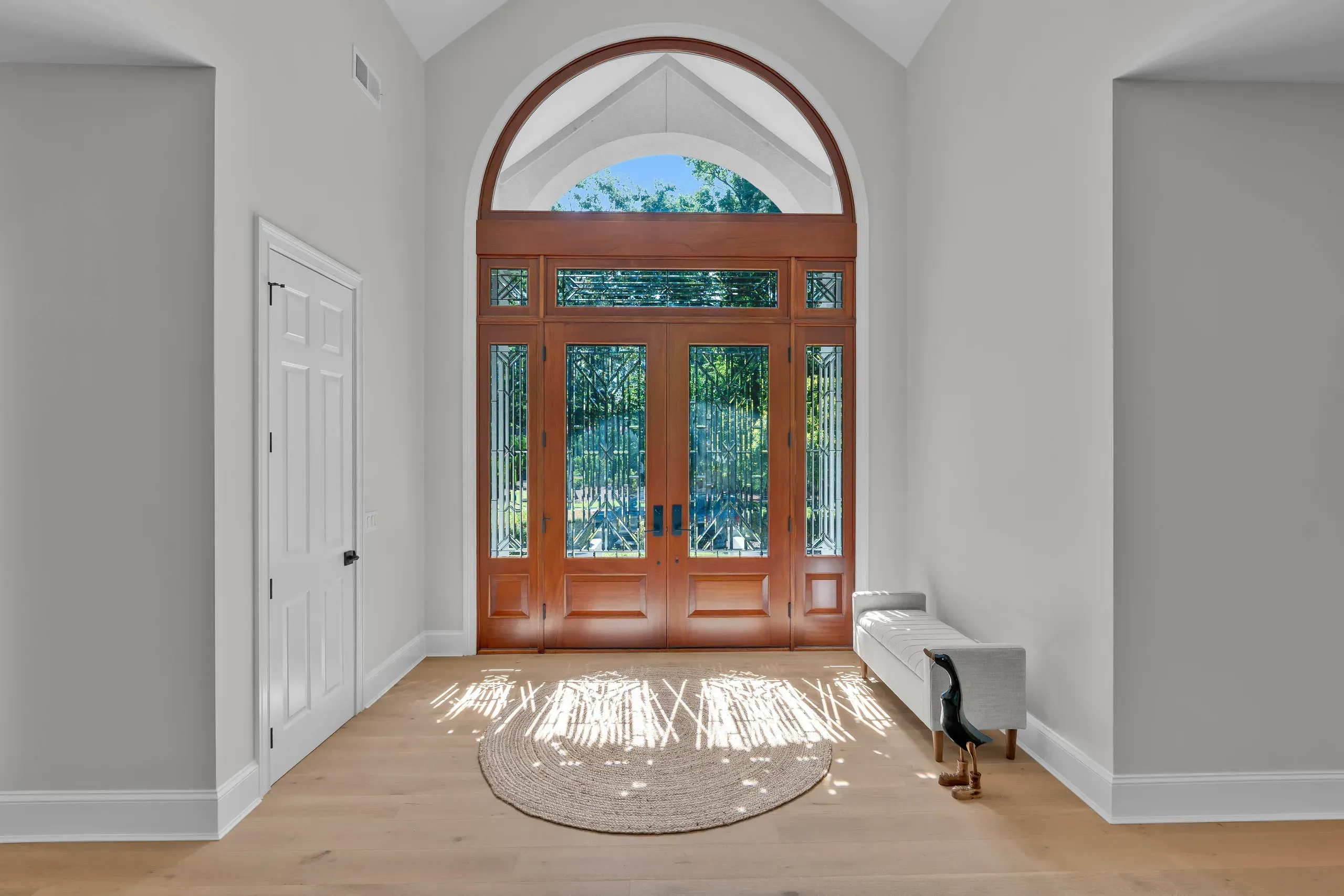 A grand foyer features tall wood double doors with an arched glass transom, decorative glass panels, light wood flooring, a round woven rug, and a cushioned bench beneath soft natural light.