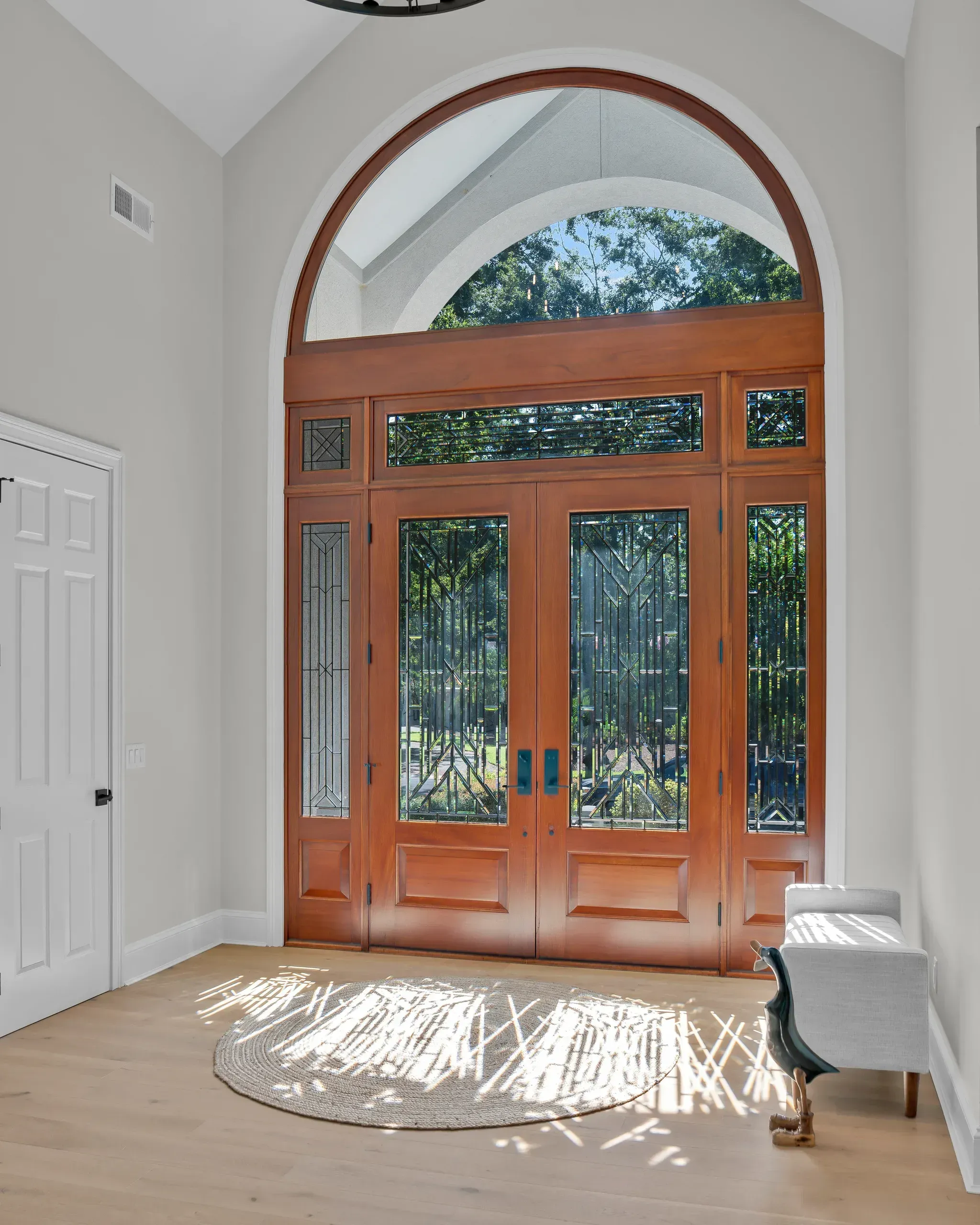 A grand foyer features a stained-glass double front door topped with a large arched transom window that fills the entryway with natural light, above light wood flooring and a round jute rug.