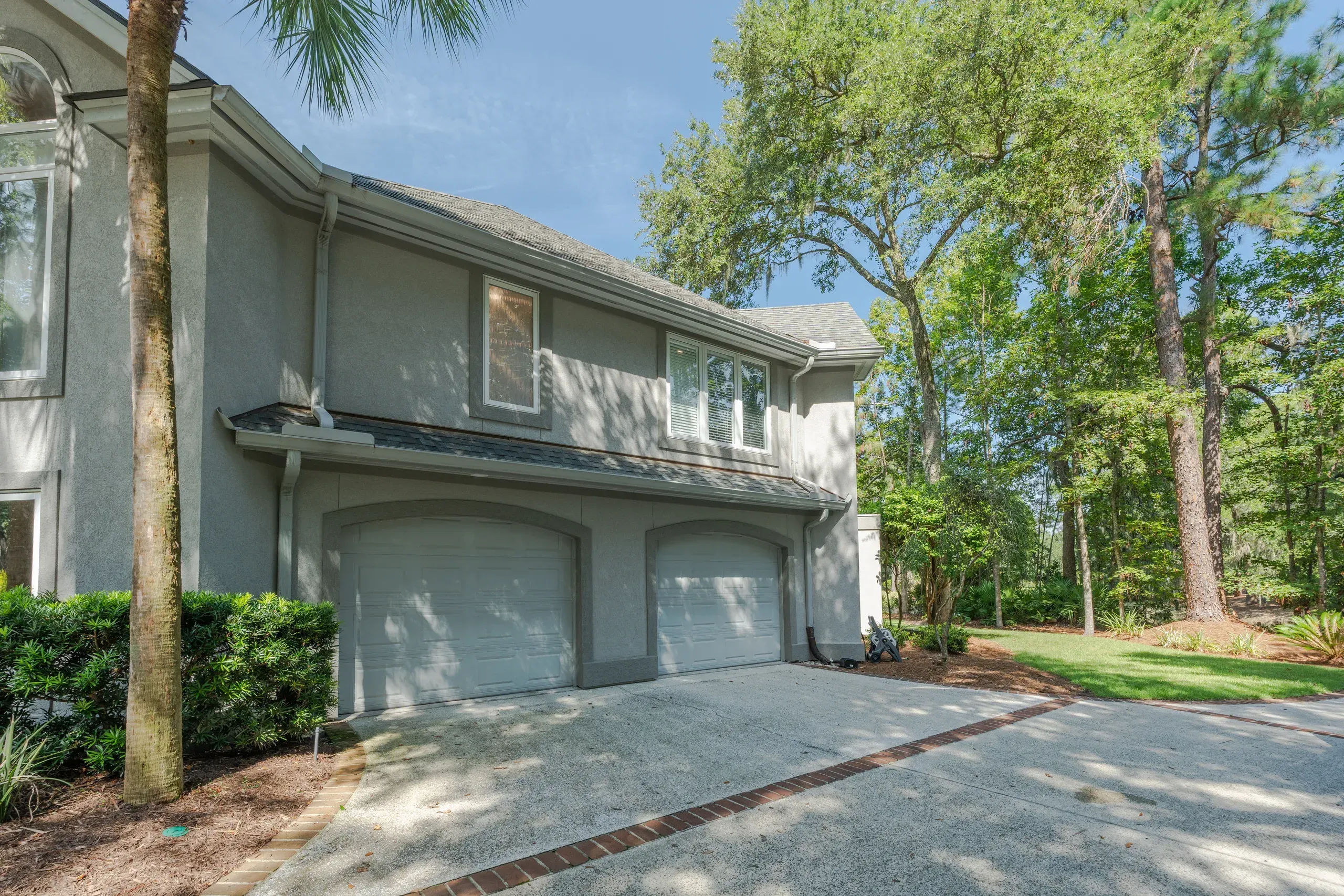 A two-story stucco home with a two-car garage sits beneath tall pine trees and a palm tree beside a concrete driveway edged in brick.