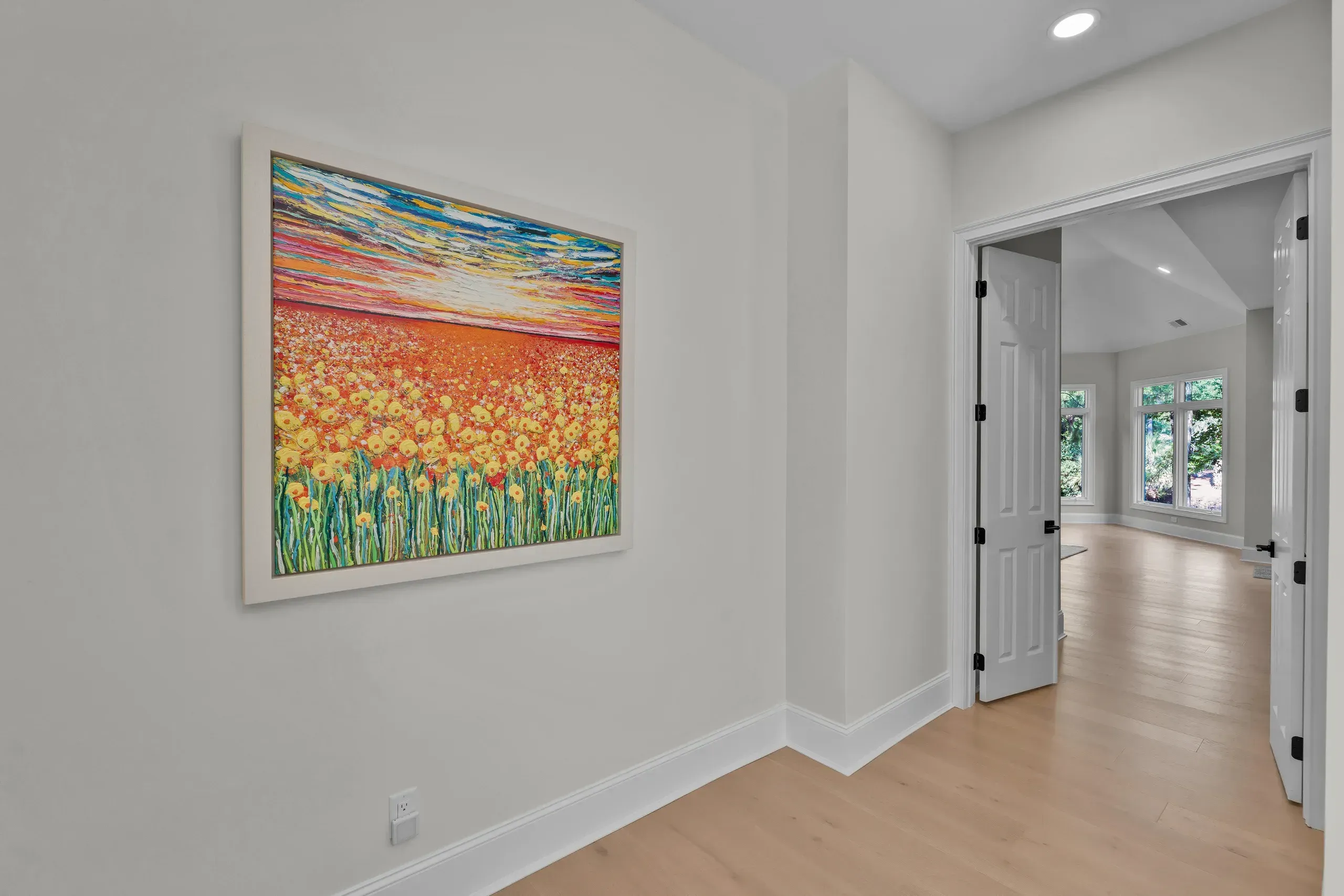 A bright hallway with light wood flooring and gray walls features a large framed floral landscape painting beside an open white door leading to a sunlit room with windows.
