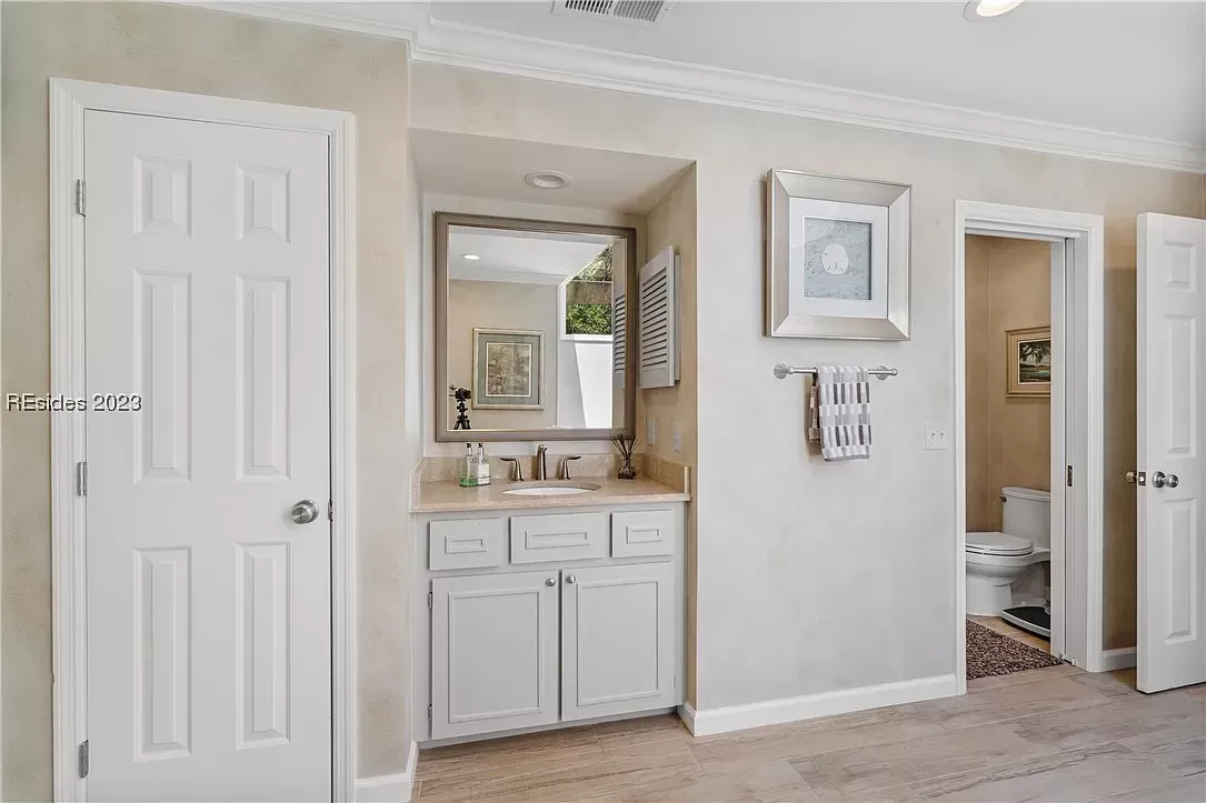 A hallway bathroom features a small vanity alcove with a framed mirror and gray cabinetry, leading past a doorway to a separate toilet room finished with warm-toned walls and wood-look flooring throughout.