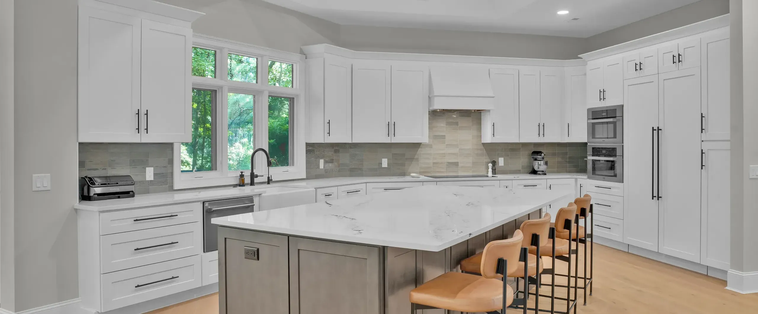 A bright kitchen remodel features white shaker cabinets, a large gray island with a white quartz countertop and leather bar stools, wood ceiling beams, and a wall of windows overlooking greenery.