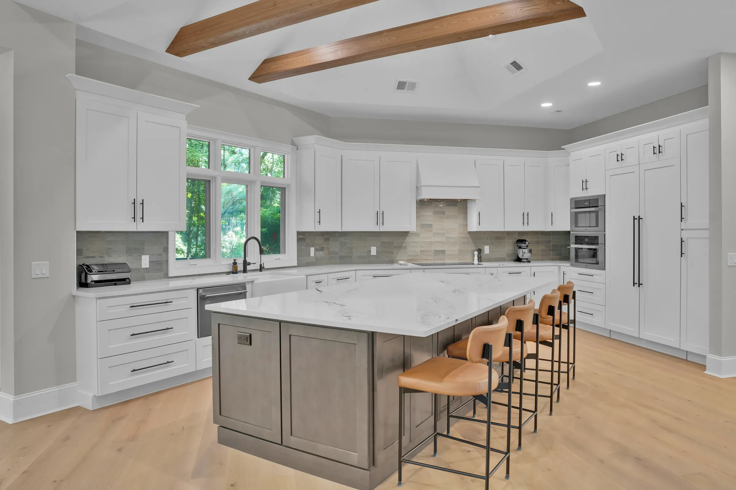 A bright kitchen remodel features white shaker cabinets, a large gray island with a white quartz countertop and leather bar stools, wood ceiling beams, and a wall of windows overlooking greenery.