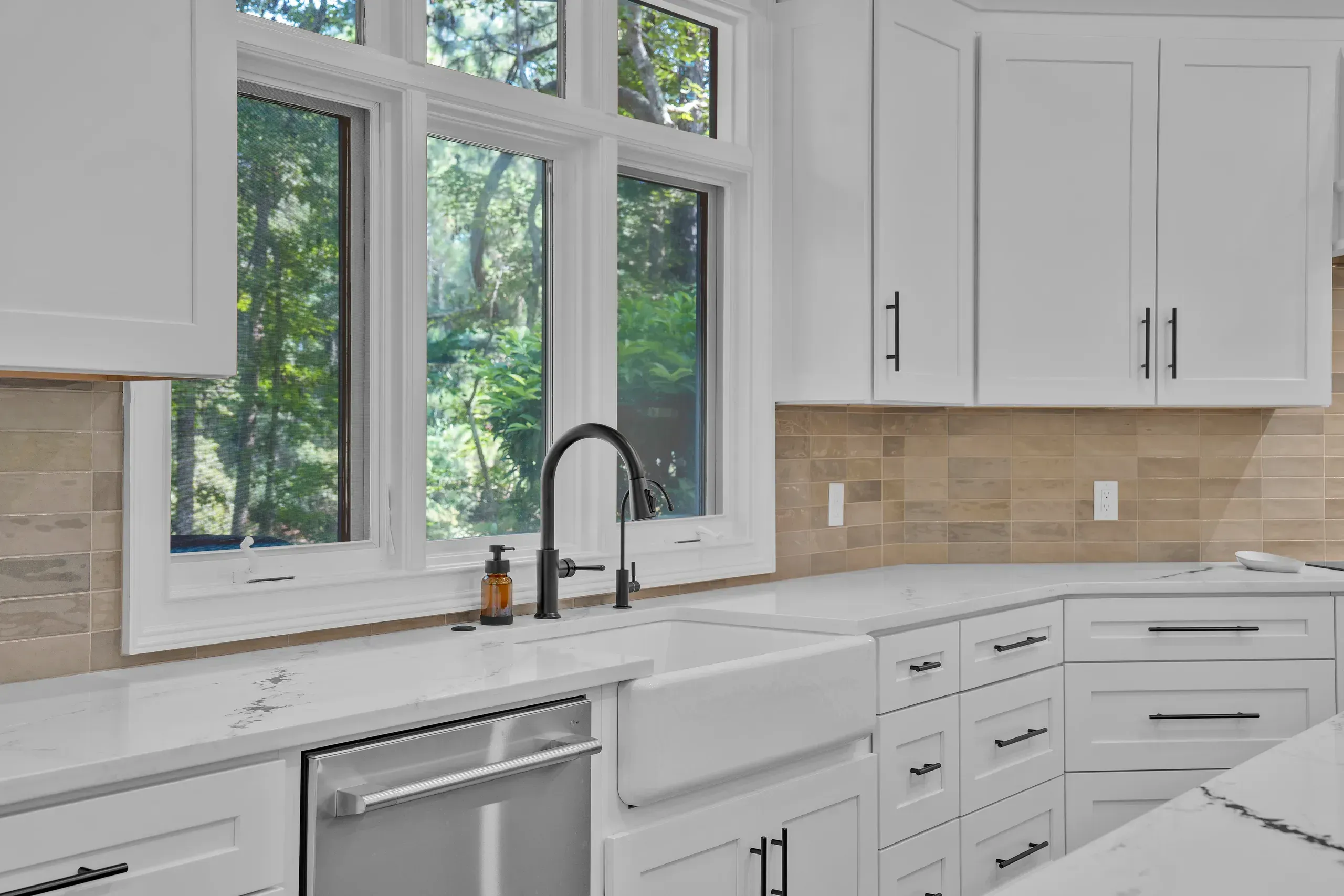 A white farmhouse sink with a matte black gooseneck faucet sits beneath a bank of large windows overlooking wooded greenery in a remodeled kitchen with white shaker cabinets and a tan tile backsplash.
