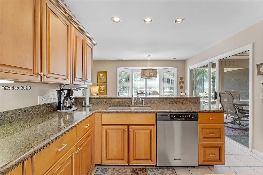 A kitchen with light wood cabinetry and granite countertops features a stainless steel dishwasher and sink beneath a bay window, opening to a sunroom with sliding glass doors leading to a patio.