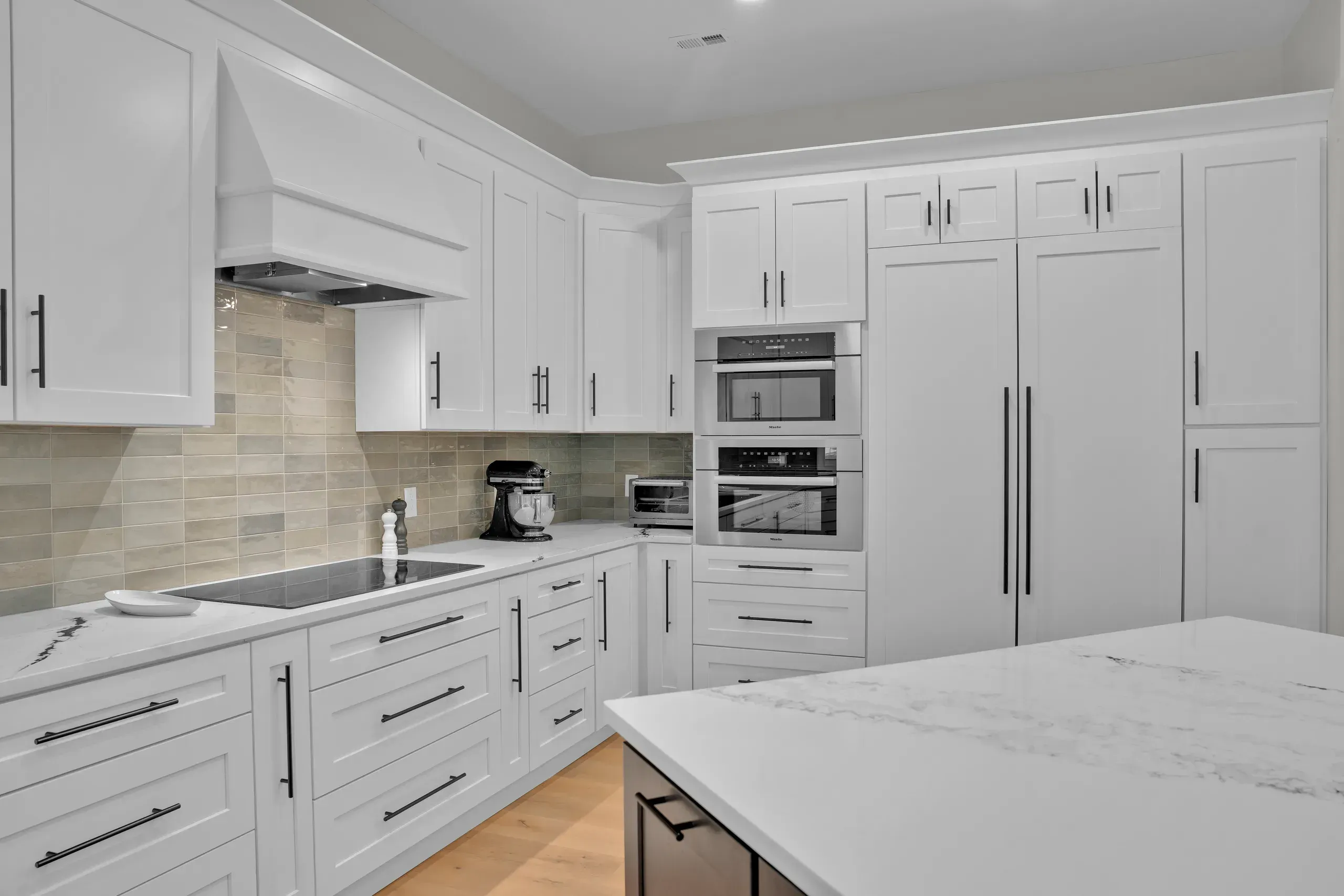 A remodeled kitchen features white shaker cabinets, a green and beige tile backsplash, a built-in cooktop, and a stainless steel double wall oven flanked by a paneled refrigerator.