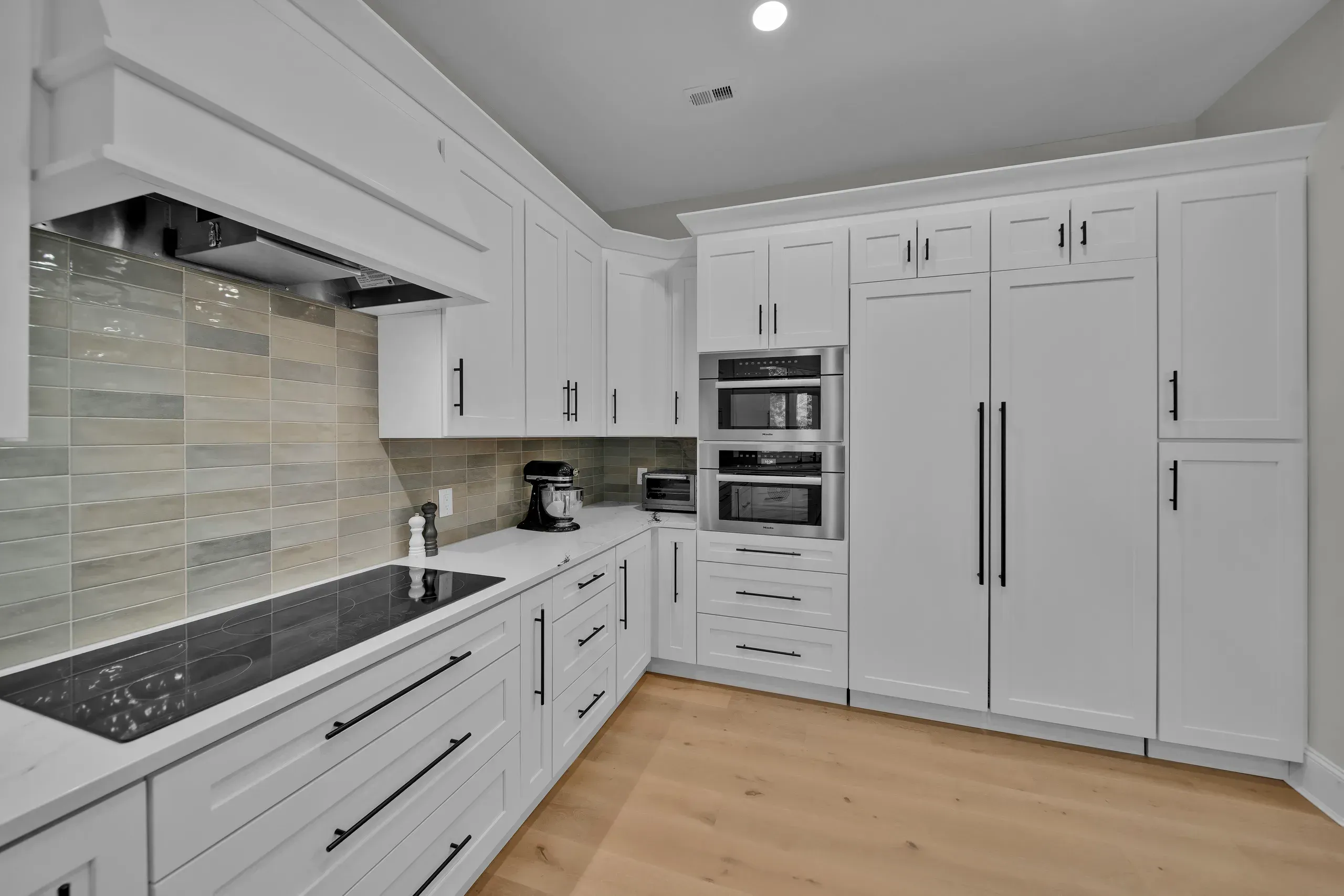A white shaker-cabinet kitchen features a built-in double oven, paneled refrigerator, cooktop with vented hood, and a multicolor tile backsplash above light wood flooring.
