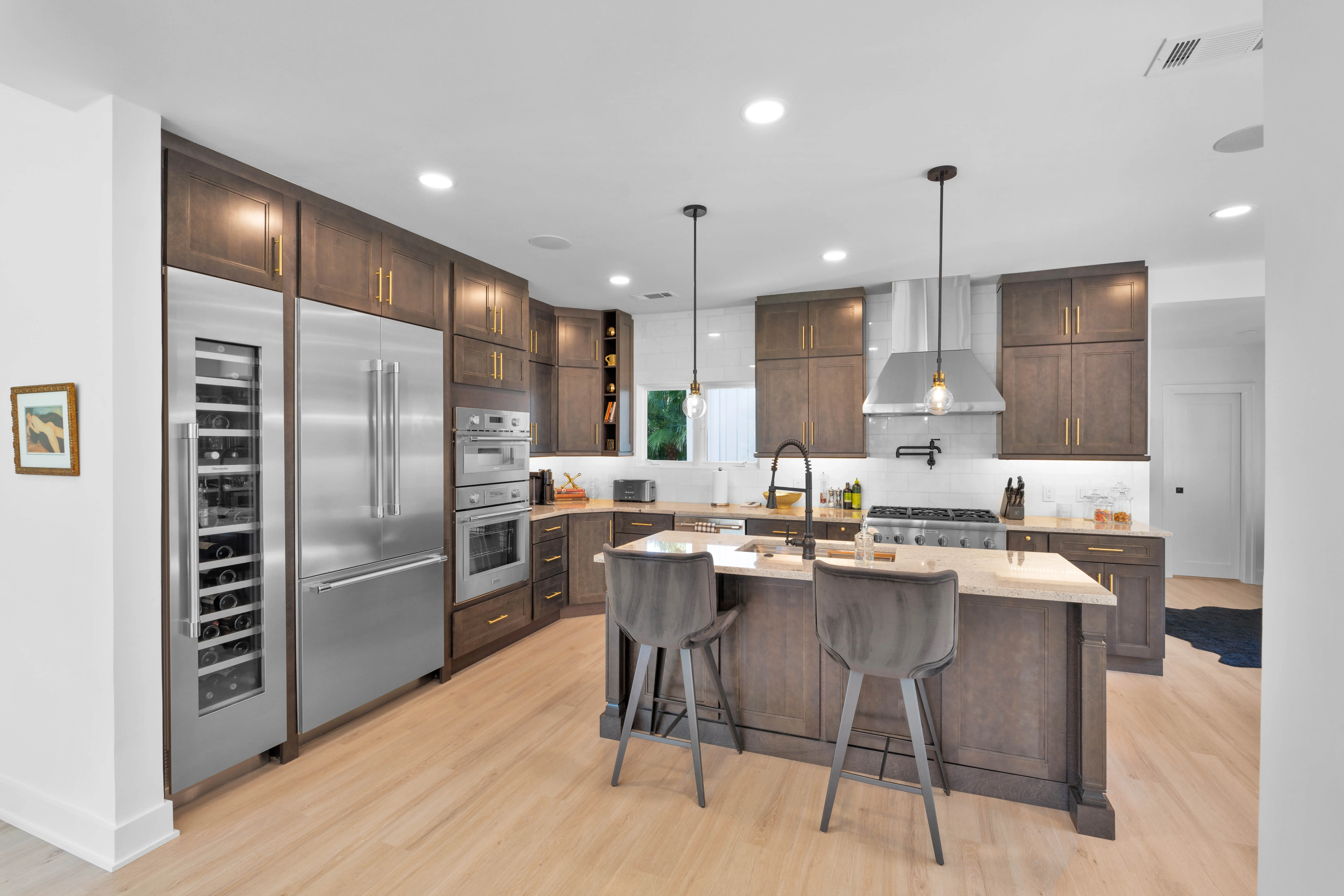 A remodeled kitchen features dark wood cabinetry, stainless steel appliances including a built-in wine refrigerator, a large center island with gray upholstered stools, and a stainless range hood over a gas cooktop.