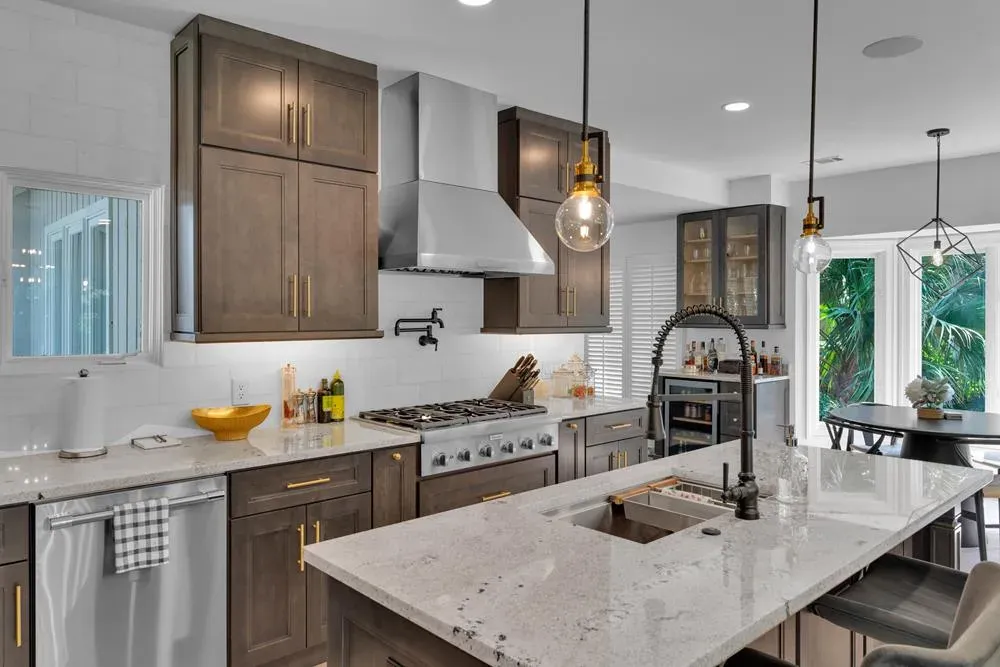 A remodeled kitchen features dark wood cabinetry with brass hardware, a stainless steel range hood over a gas cooktop, and a large granite island with a black gooseneck faucet, opening onto a bright breakfast nook with a view of palm trees outside.