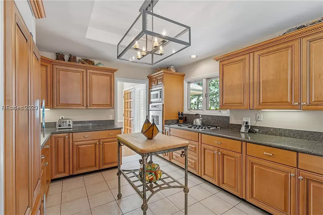A kitchen with rich wood cabinetry, dark granite countertops, a tile floor, and a small metal-framed island beneath a geometric candle chandelier.