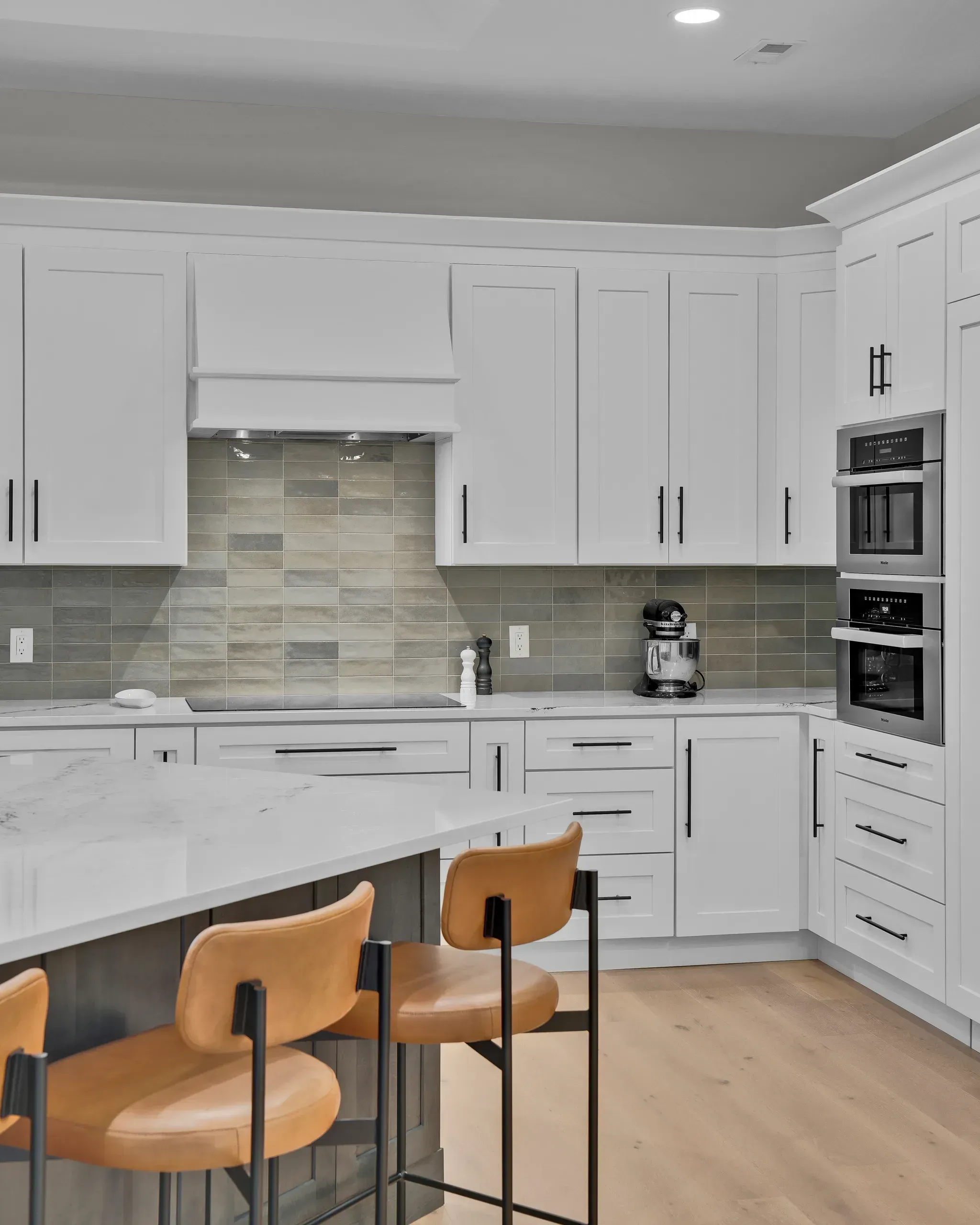 A bright white kitchen features a tiled cooktop backsplash, stainless double wall ovens, a marble-look island with tan leather bar stools, and light hardwood flooring.