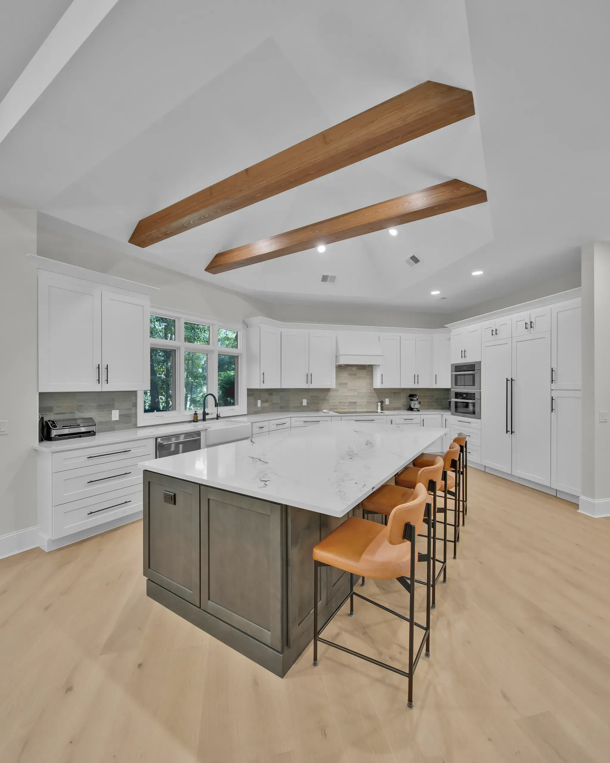 A vaulted white kitchen ceiling with two exposed wood beams sits above a large marble-topped island lined with orange leather barstools, white shaker cabinetry, and light hardwood flooring.