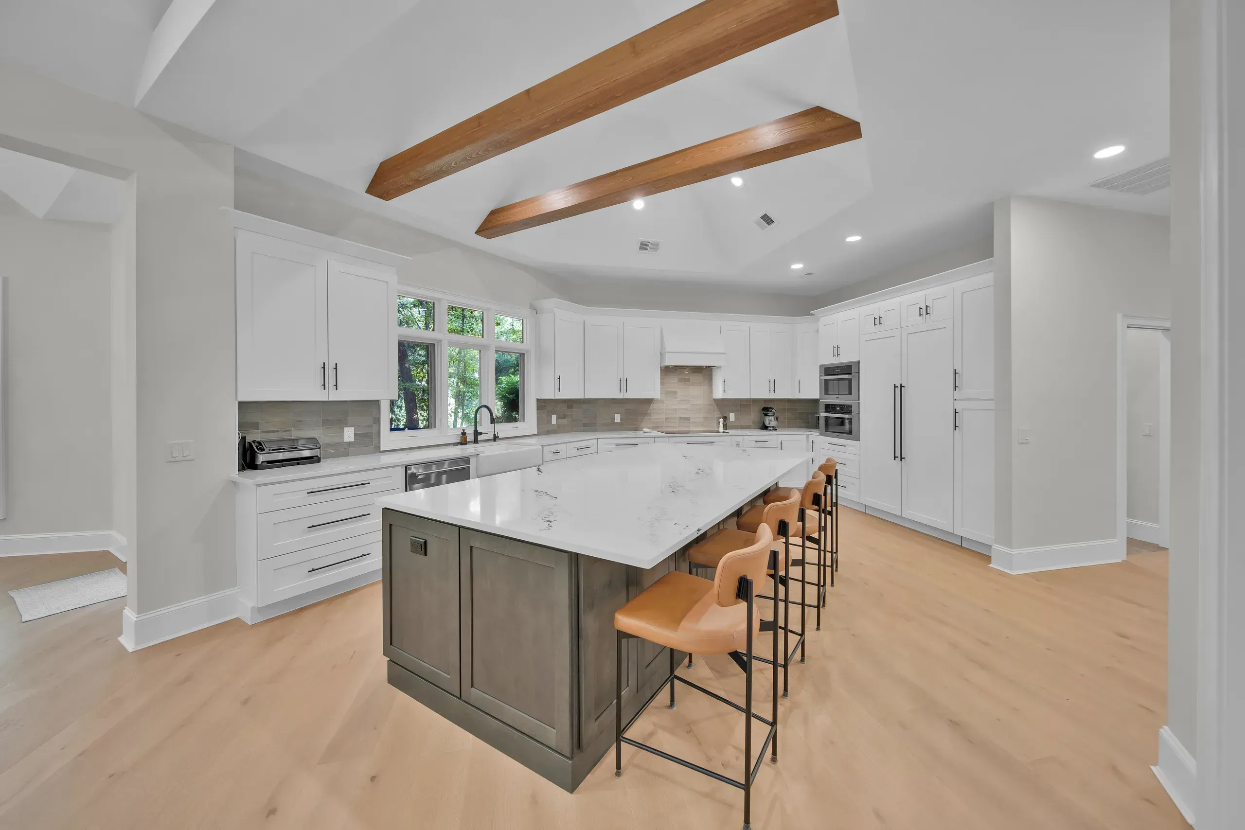 A vaulted kitchen with exposed wood ceiling beams features a large gray island topped with white quartz and surrounded by tan leather counter stools, alongside white cabinetry and stainless steel appliances.