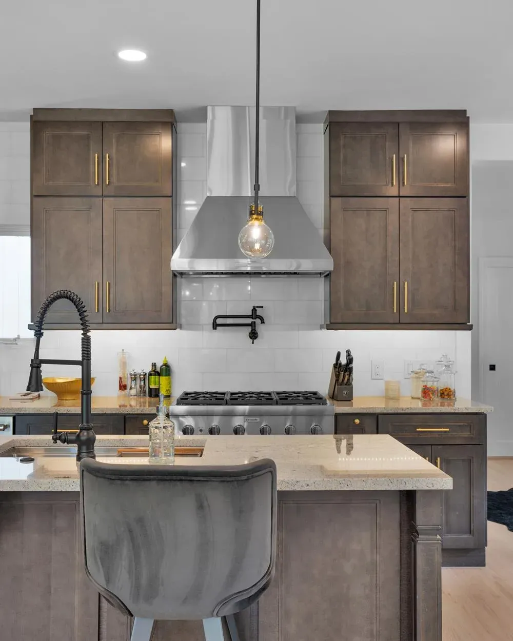 A kitchen remodel features dark wood cabinetry with brass hardware, a stainless steel range and hood with pot filler, quartz countertops, and a large island with a gray upholstered stool.