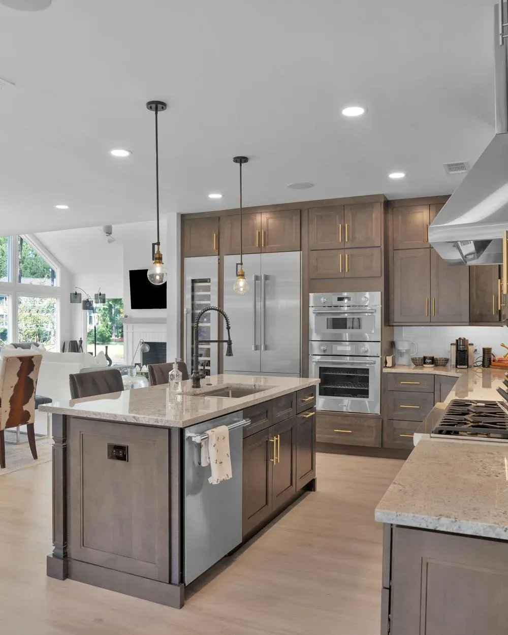 An open-concept kitchen remodel features a large island with a farmhouse sink, gray-toned wood cabinetry with gold hardware, stainless steel built-in refrigerator and double ovens, and a view into a bright living area with vaulted ceilings.