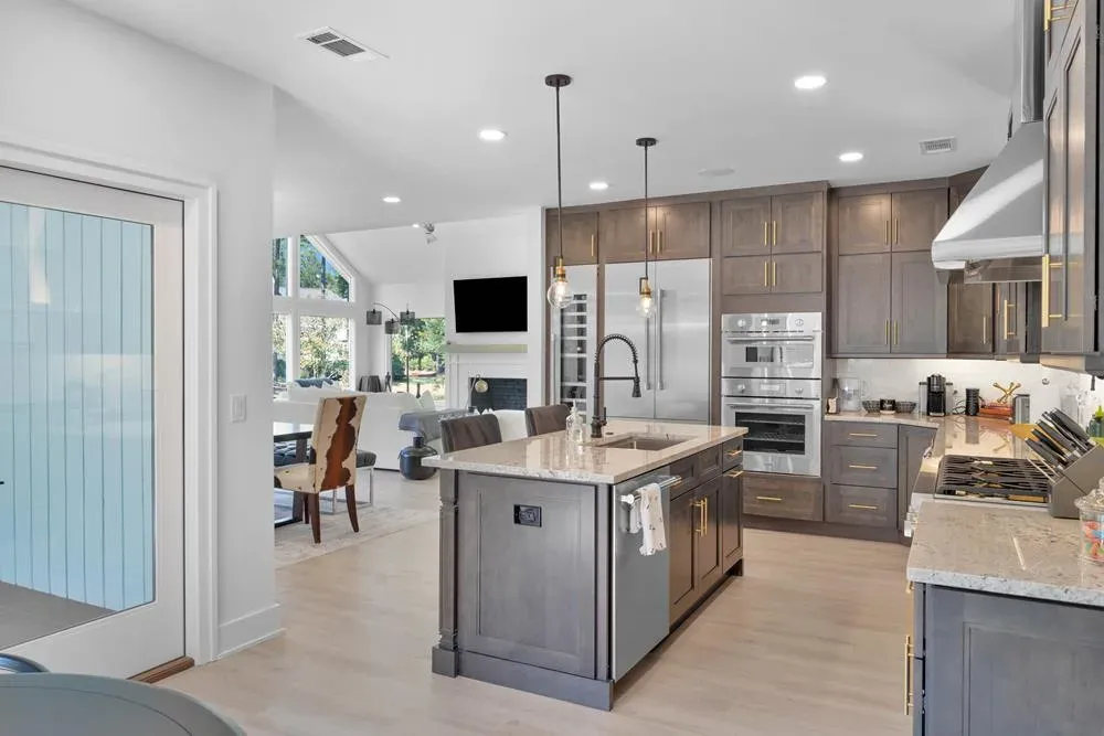 An open-concept kitchen features a large center island with a farmhouse sink, dark wood cabinetry with brass hardware, and stainless steel built-in appliances, opening into a bright vaulted-ceiling living area beyond.