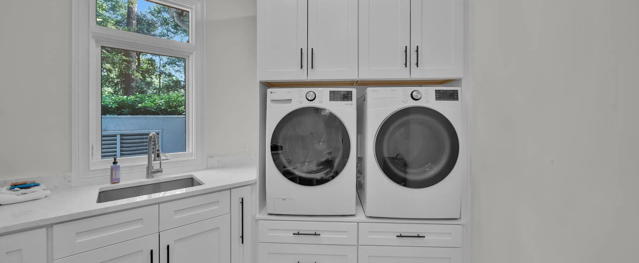 A bright laundry room remodel features white front-load washer and dryer set beneath white upper cabinets, alongside a farmhouse-style sink with quartz countertops and a window overlooking green trees.
