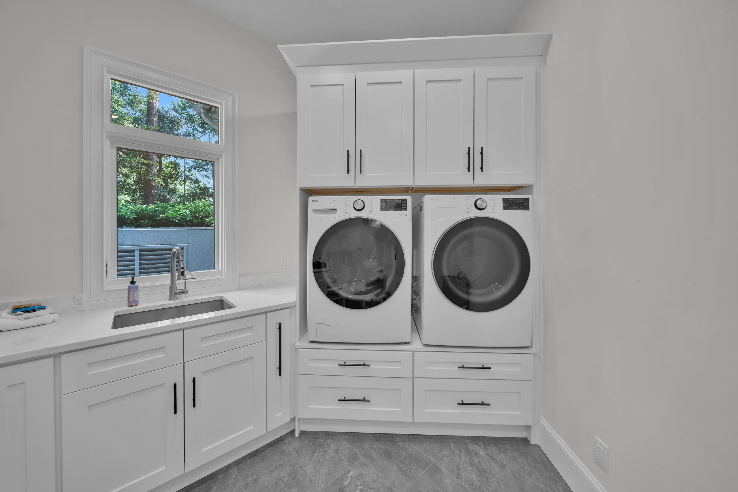 A bright laundry room remodel features white front-load washer and dryer set beneath white upper cabinets, alongside a farmhouse-style sink with quartz countertops and a window overlooking green trees.