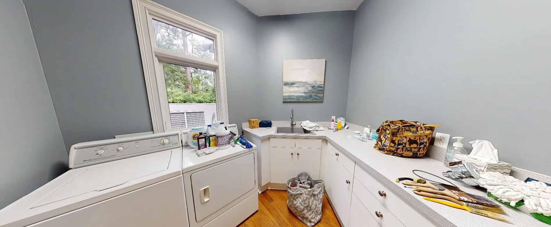 A laundry room with gray walls features a white washer and dryer beside a wraparound counter with a built-in sink, white cabinetry, and a large window overlooking greenery.