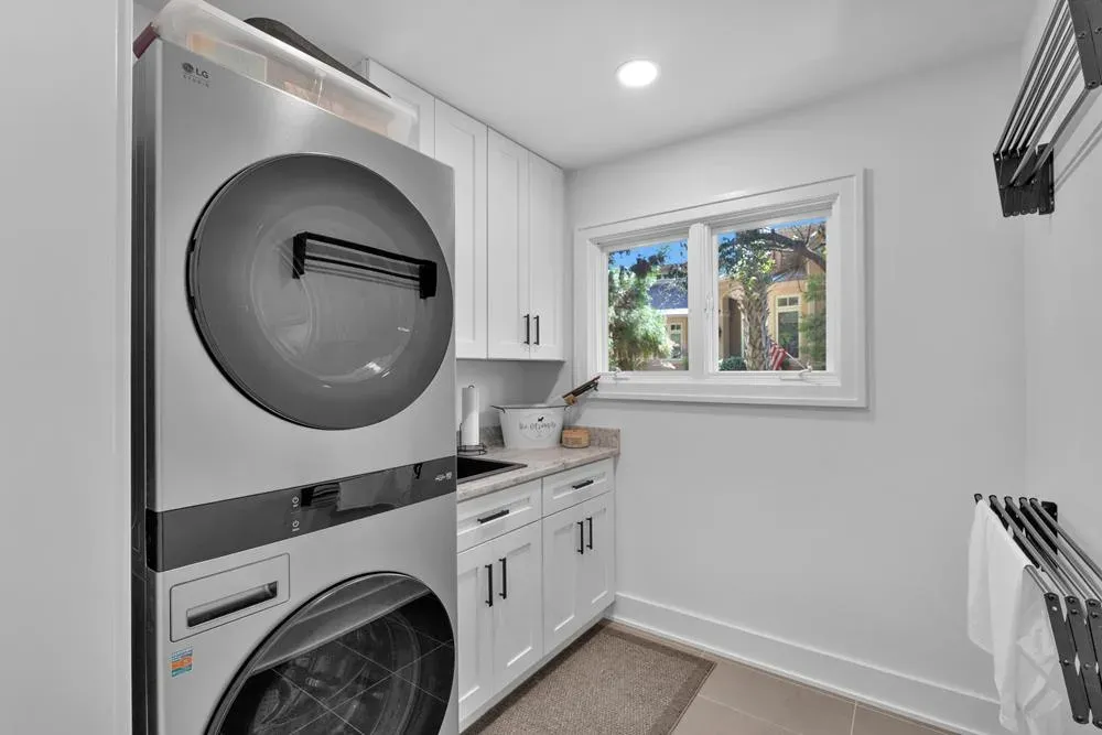 A bright laundry room features a stacked stainless steel washer and dryer beside white cabinetry, a small utility sink, and a window with a view of trees and a nearby home.
