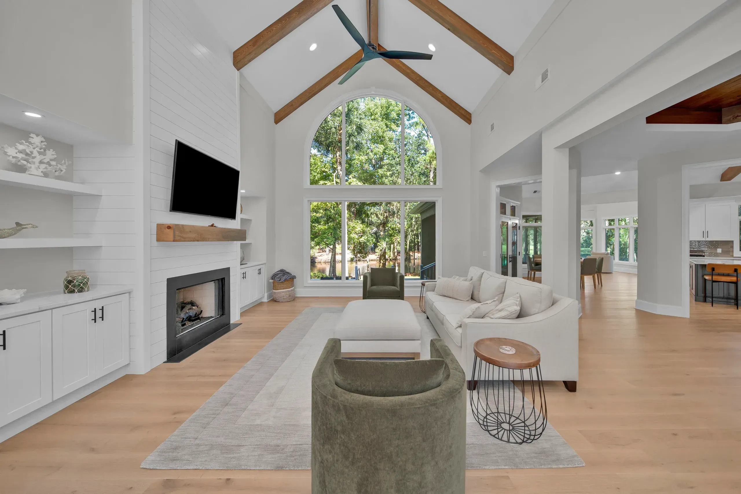 A great room with vaulted ceilings and exposed wood beams features a shiplap fireplace wall, a large arched window overlooking wooded scenery, and light hardwood floors with neutral seating.