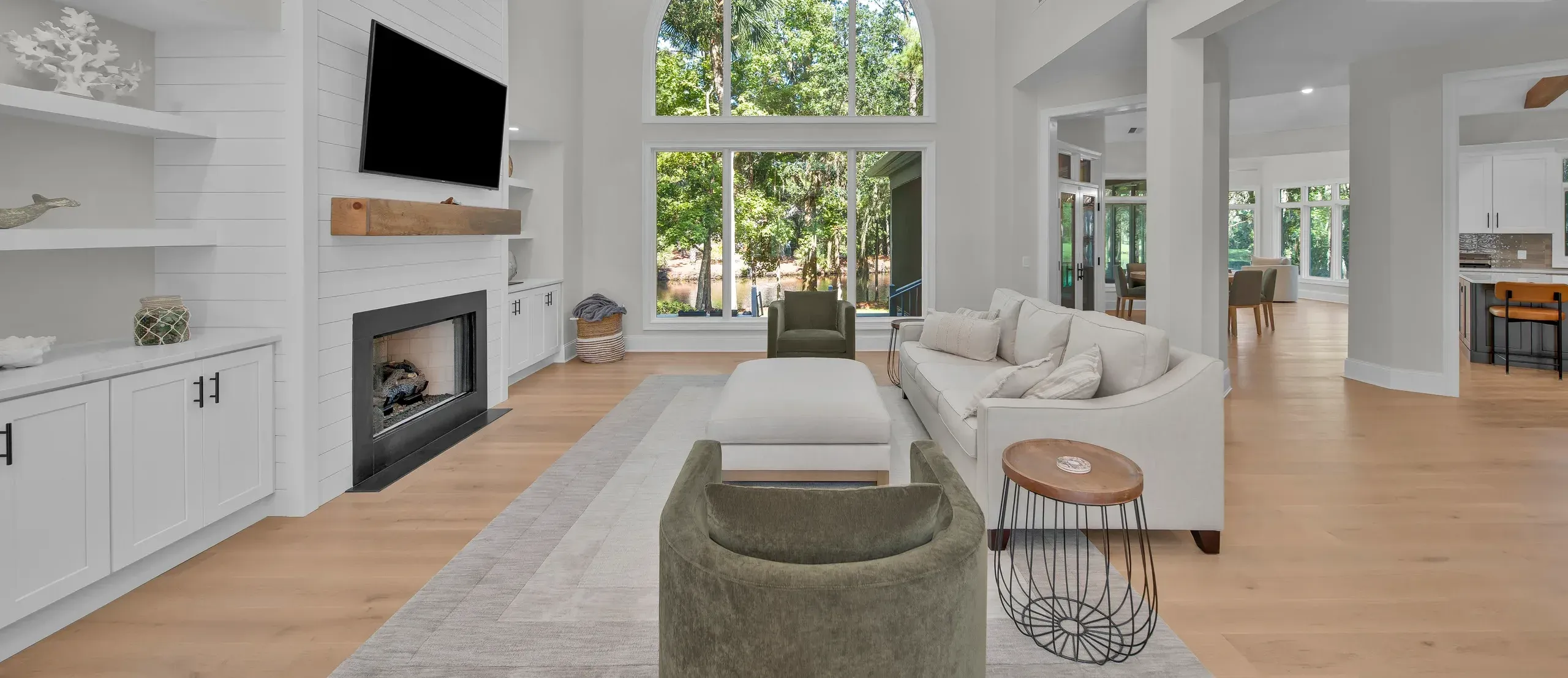 A great room with vaulted ceilings and exposed wood beams features a shiplap fireplace wall, a large arched window overlooking wooded scenery, and light hardwood floors with neutral seating.