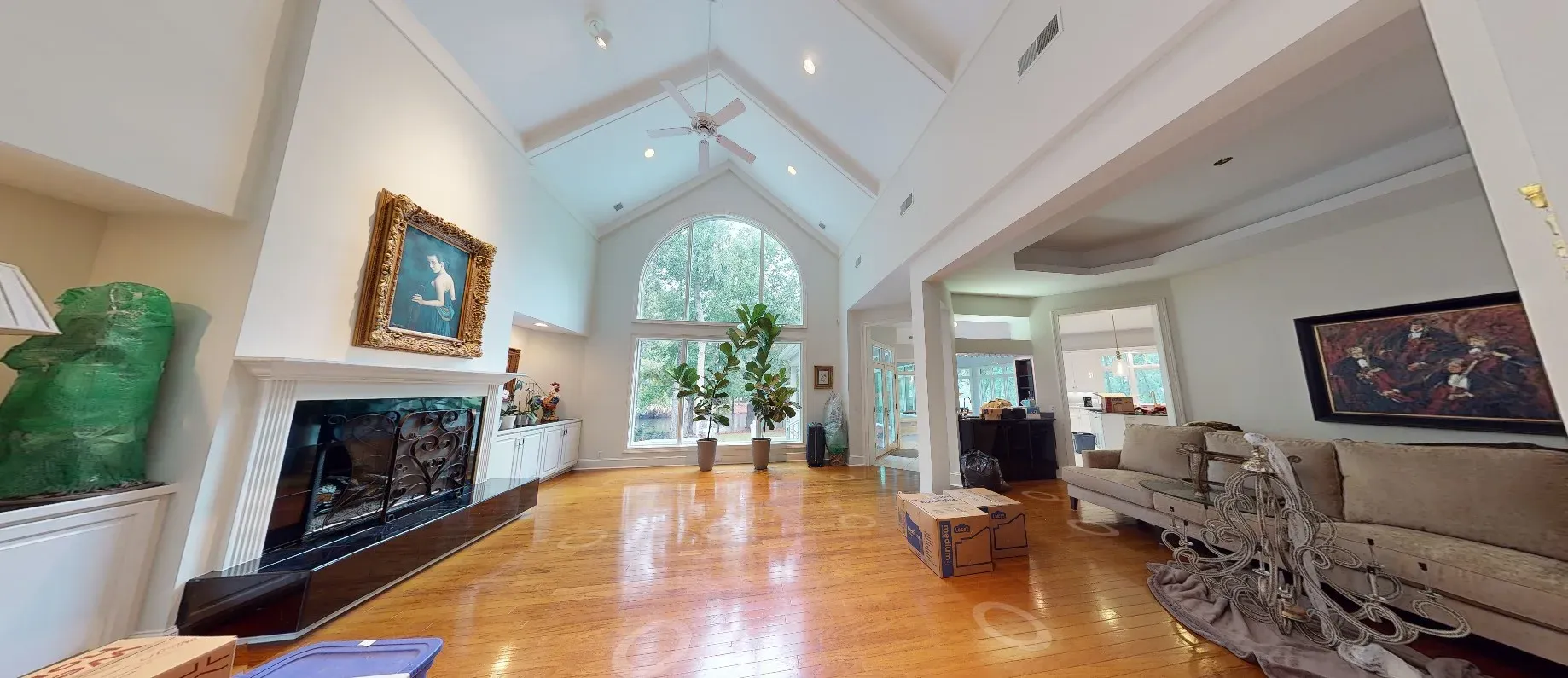 A spacious living room with vaulted ceilings, exposed beams, a large arched window, hardwood floors, and a fireplace with a decorative iron screen, currently mid-move with boxes on the floor.