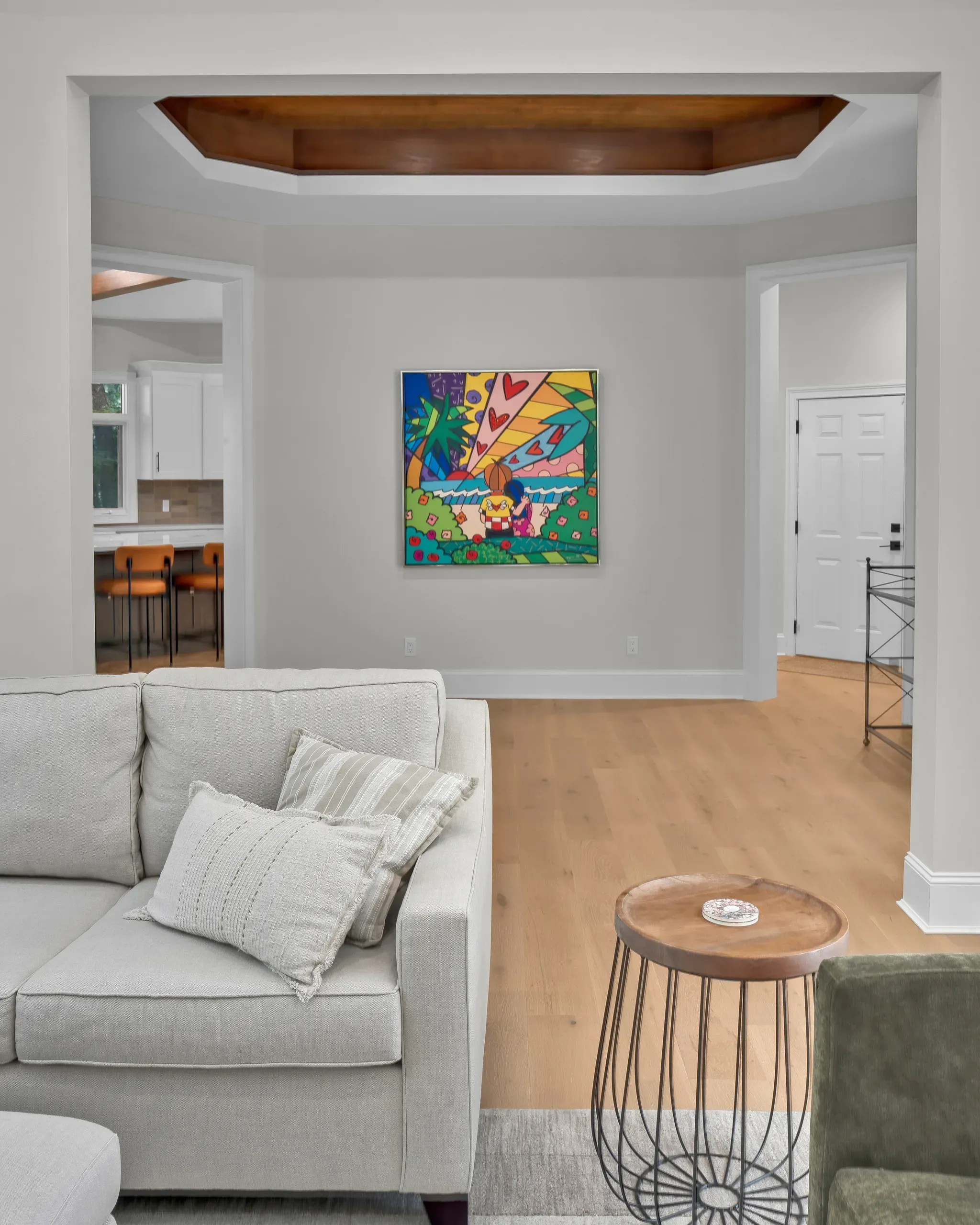 A bright living room with a light gray sectional sofa and wood side table opens through wide doorways to a white kitchen and an entry foyer, with wood flooring throughout and a colorful framed painting on the wall beneath a wood-paneled tray ceiling.