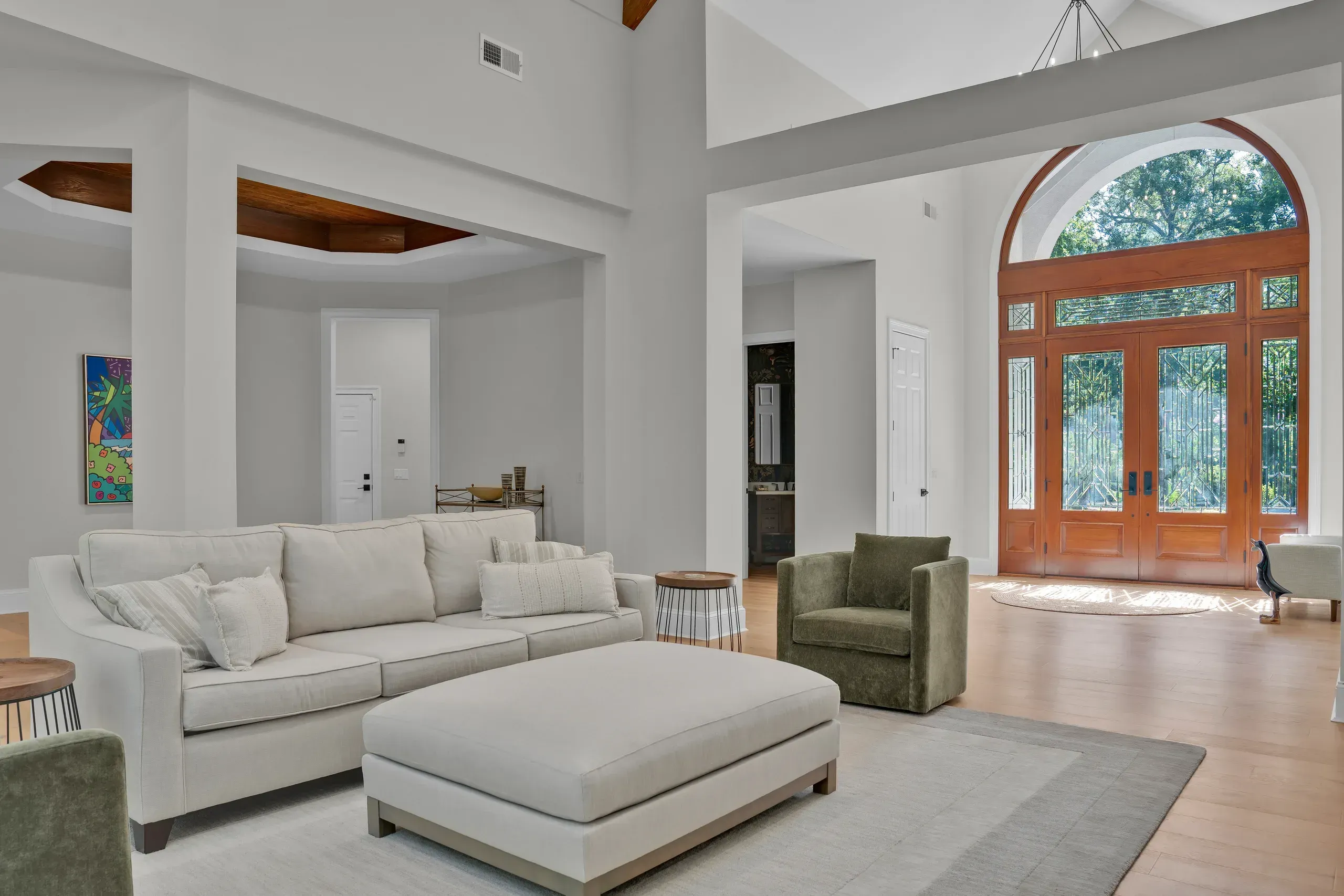 An open-concept living room with a light gray sectional sofa, ottoman, and green velvet armchairs faces a grand foyer featuring an arched wooden double door with stained glass and a view of trees outside.
