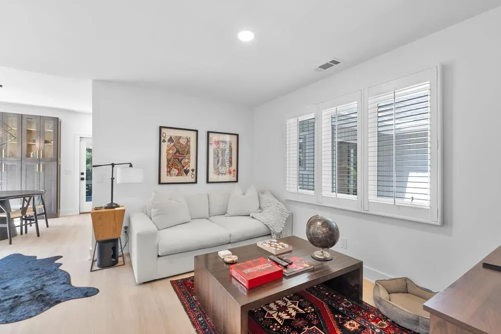 A bright living room features a light gray sofa, a wooden coffee table with a patterned rug beneath it, and large windows fitted with white plantation shutters overlooking greenery outside.