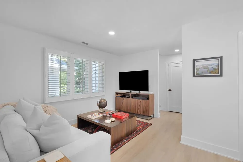 A bright living room features a white sofa, wooden coffee table with a patterned rug, a media console with a mounted television, plantation shutters on the window, and light hardwood flooring leading to a hallway.