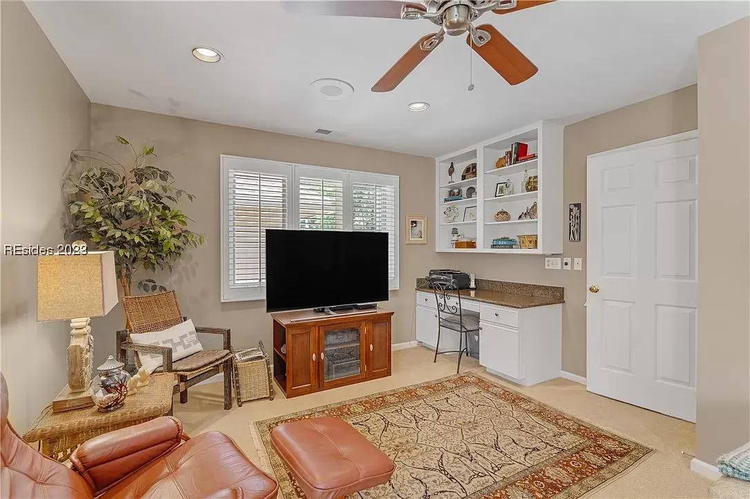 A cozy living room with a wooden TV console, wicker chairs, a leather recliner and ottoman on an area rug, plantation shutters over a window, and a built-in desk with shelving beside the entry door.