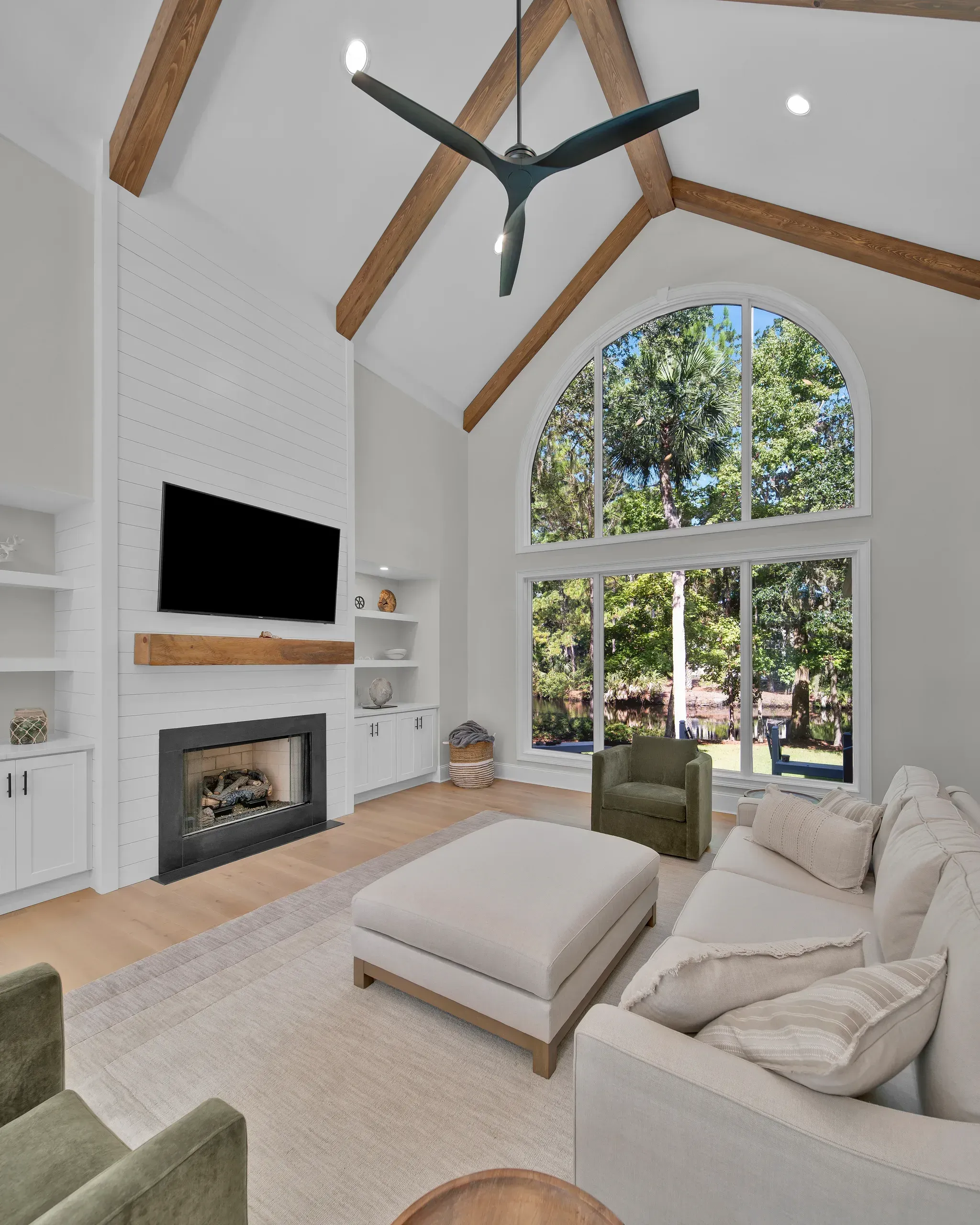 A vaulted living room with exposed wood ceiling beams features a shiplap fireplace wall, built-in cabinetry, and a two-story arched window overlooking wooded grounds and a lagoon.