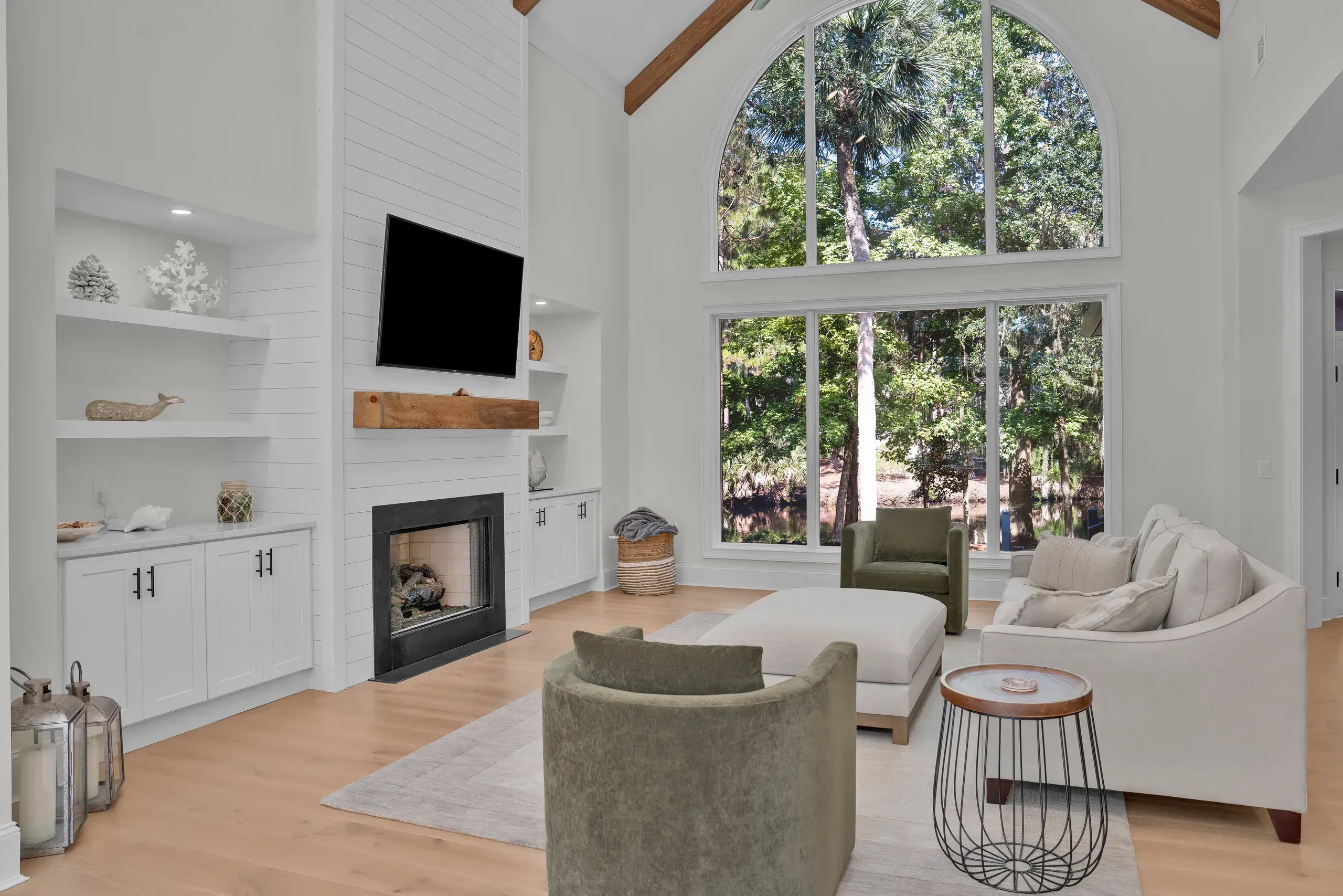 A vaulted living room with exposed wood beams features a shiplap fireplace surround flanked by white built-in shelving, a wall-mounted television, and a large arched window overlooking wooded views, furnished with a beige sofa and olive green armchairs on a light area rug.
