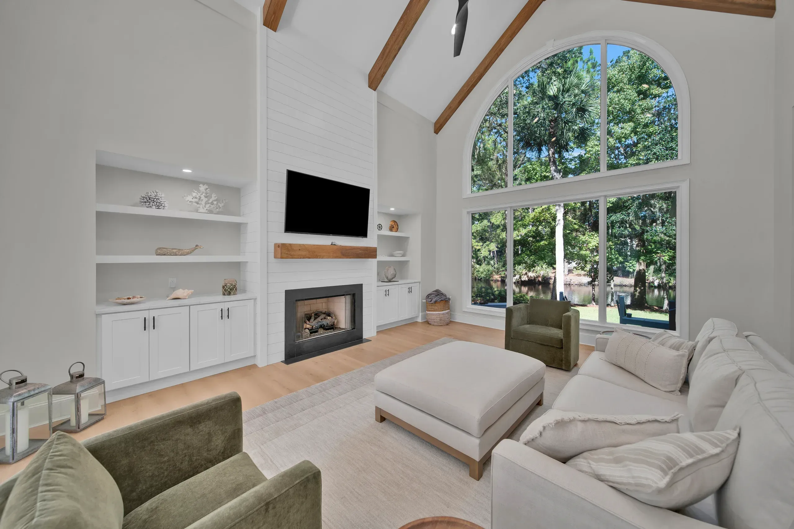 A vaulted living room with exposed wood beams features a shiplap fireplace wall flanked by built-in shelving, and a large arched window overlooks a wooded lagoon view.