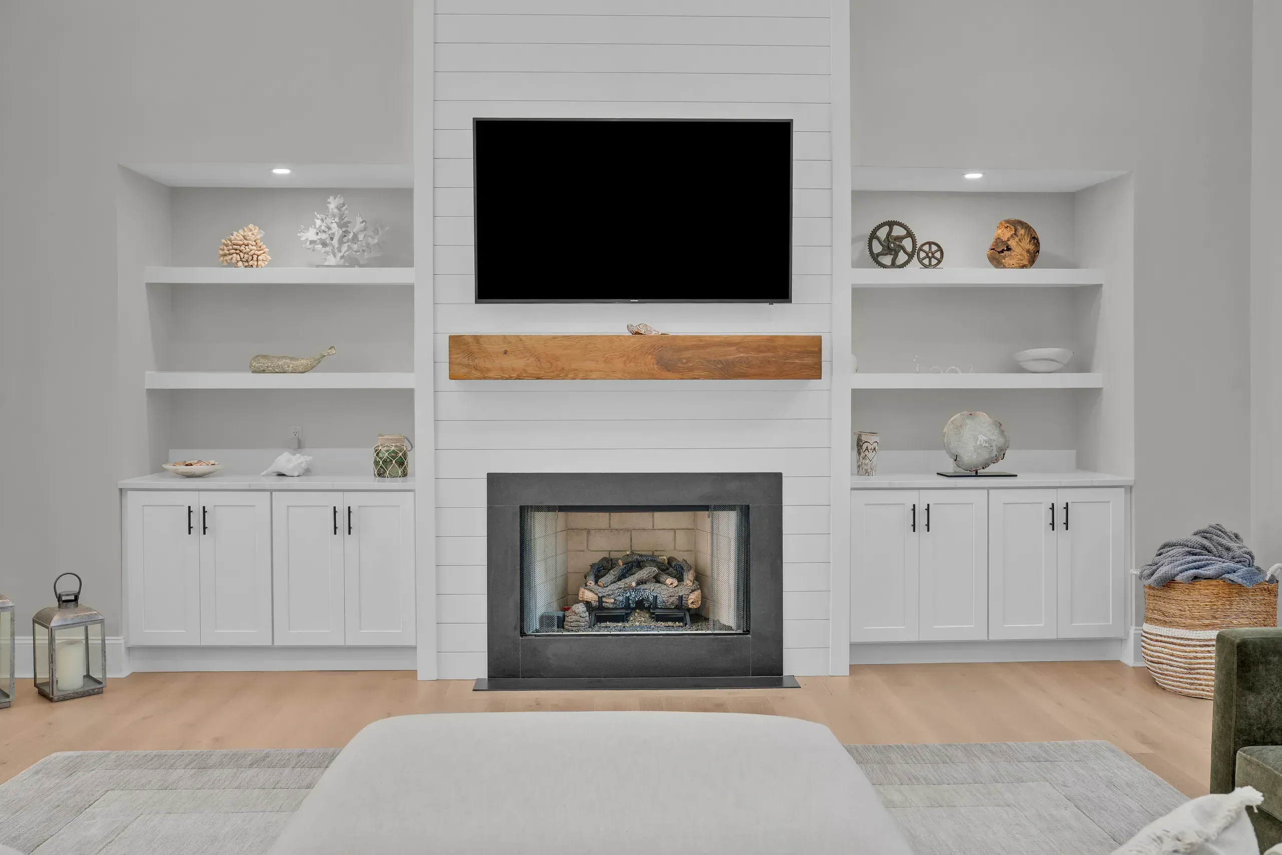 A living room features a gas fireplace framed by white shiplap paneling with a wood mantel, a wall-mounted television, and built-in cabinetry with open shelving on either side.
