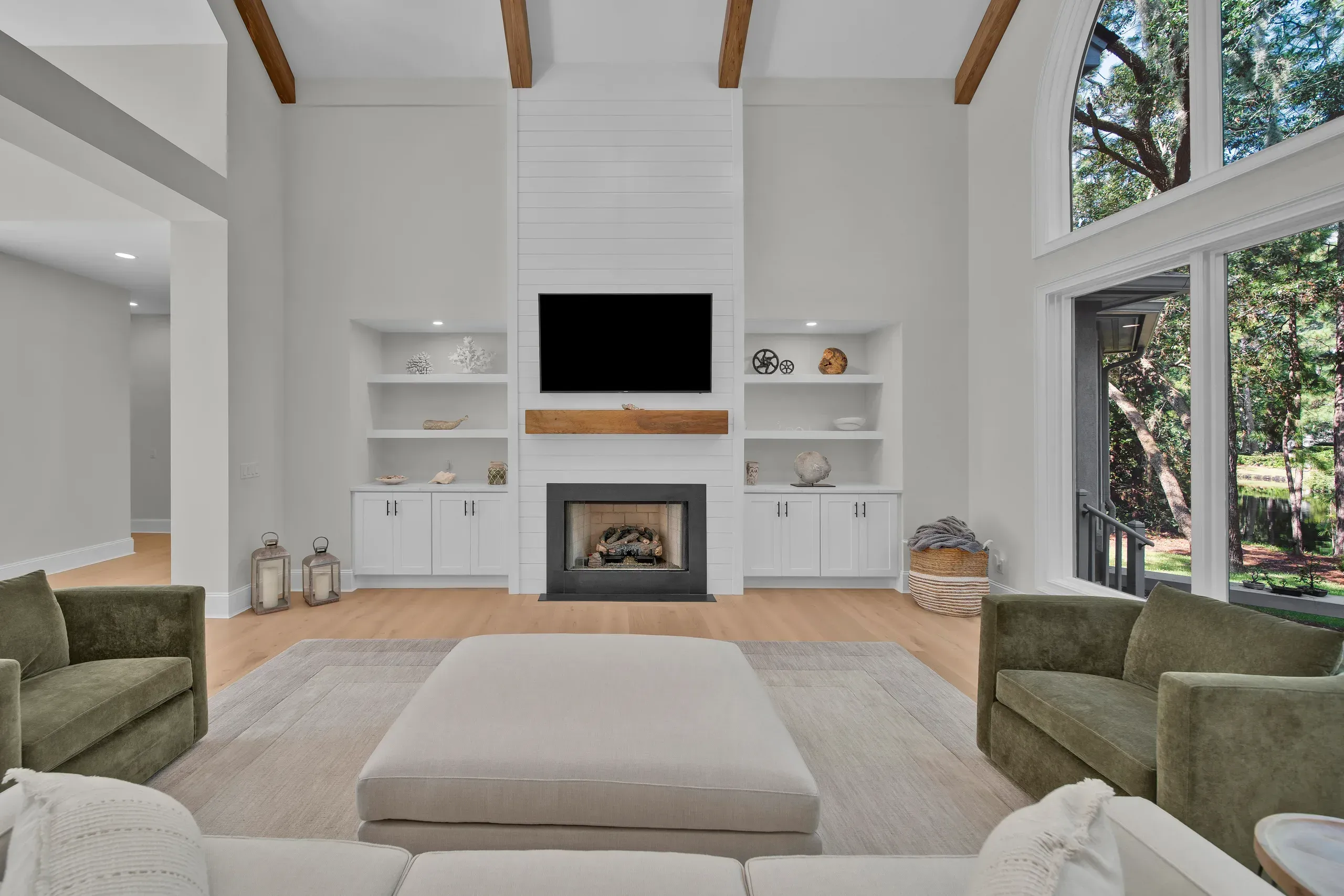 A vaulted living room features a white shiplap fireplace wall with a mounted television and wood mantel, flanked by built-in shelving, with green velvet chairs and large windows overlooking trees.