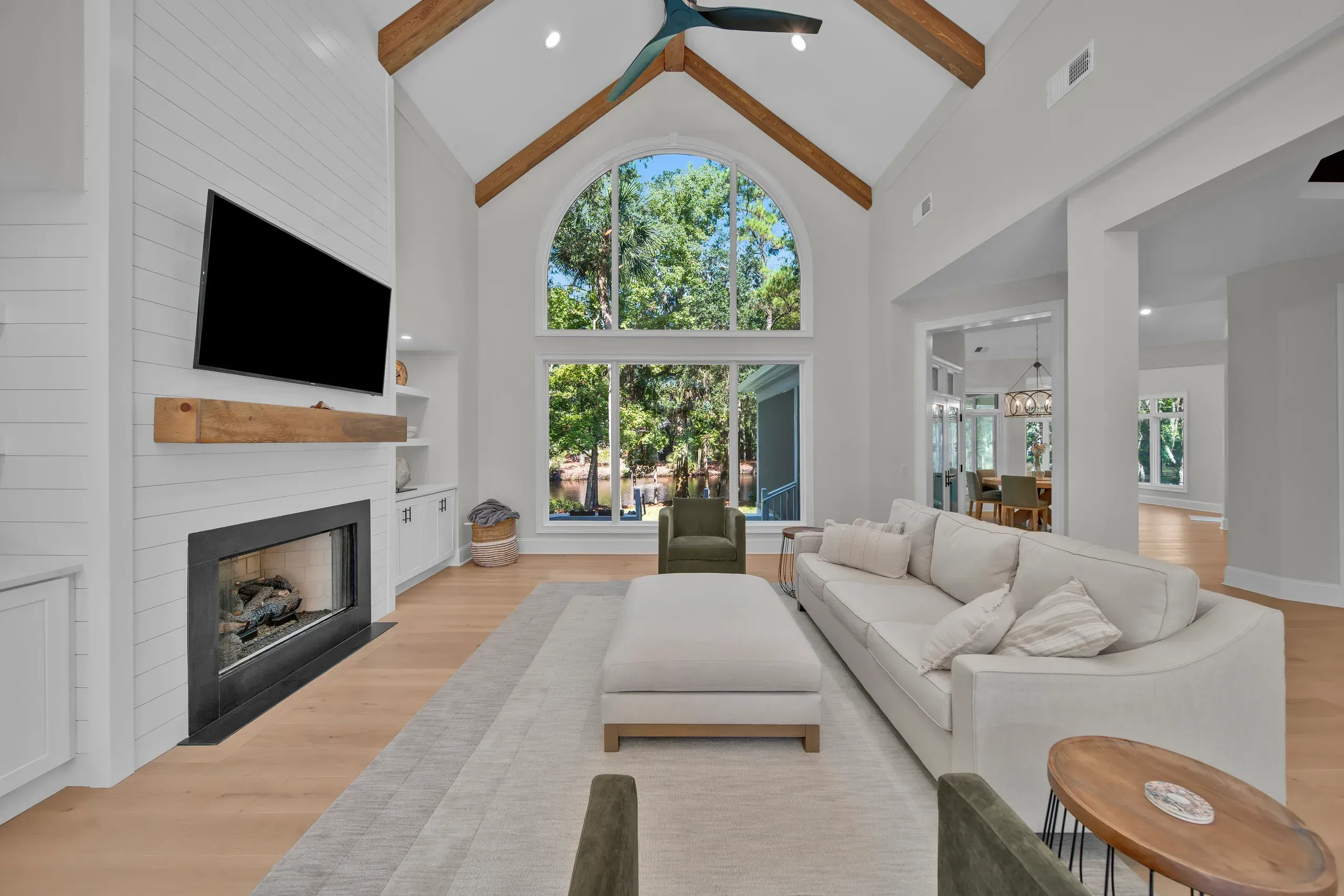 A vaulted living room with exposed wood beams and a two-story arched window overlooking wooded grounds features a shiplap fireplace wall, mounted television, and light upholstered seating on a pale rug.