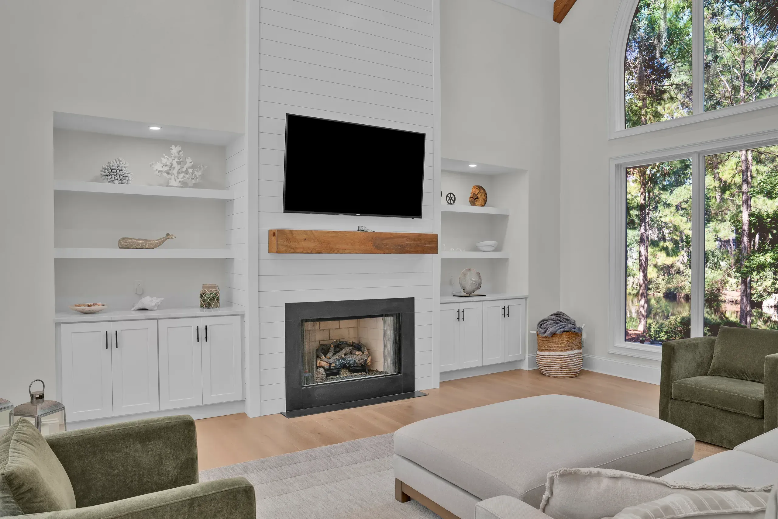 A vaulted living room features a white shiplap fireplace wall with a wood mantel and mounted television, flanked by built-in white cabinetry and shelving, with tall arched windows overlooking wooded grounds.