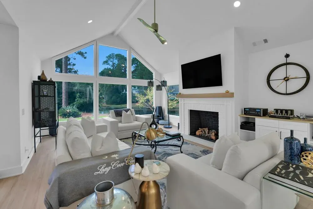 A vaulted living room features a wall of tall windows overlooking trees, a white fireplace with mounted television, and light upholstered sofas arranged around a glass coffee table.