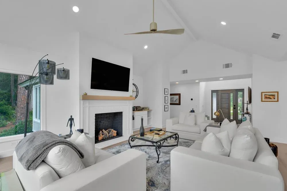A bright living room with vaulted ceilings features white sectional sofas arranged around a fireplace with a wall-mounted television, opening to an entryway with a wooden front door.