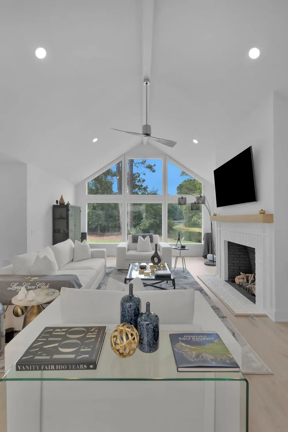 A vaulted living room with white sectional seating and a glass coffee table centers on a white brick fireplace beneath a mounted television, with tall peaked windows framing a wooded, waterside view.