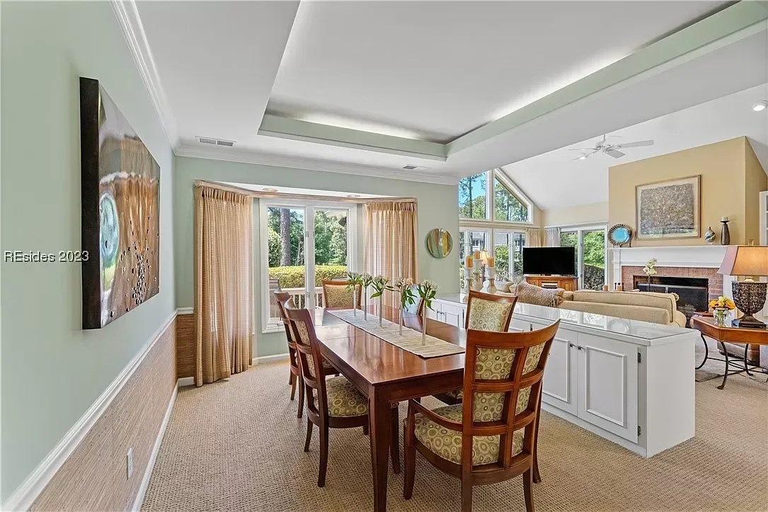 A dining area with a wooden table and patterned chairs opens into a vaulted-ceiling living room with a brick fireplace and large windows overlooking trees.