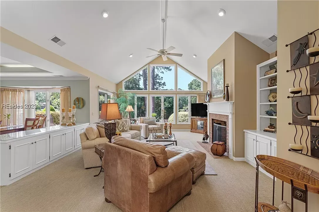 A spacious living room with vaulted ceilings and tall windows opens to built-in cabinetry, a brick fireplace, and a carpeted seating area with leather and upholstered furniture.