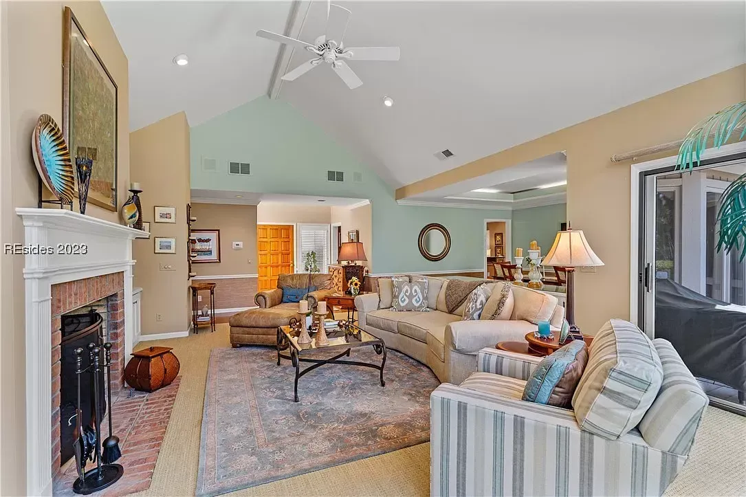 A vaulted living room with a brick fireplace, ceiling fan, and beige sectional and striped sofas arranged around a patterned area rug, opening to a dining area and sliding glass doors.